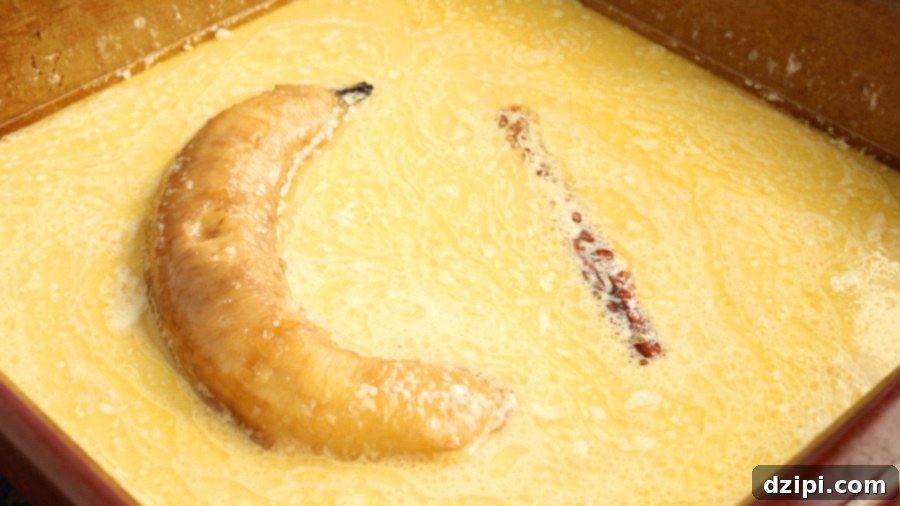 A peeled banana and a cinnamon stick are bathing in a baking dish filled with butter and cream.