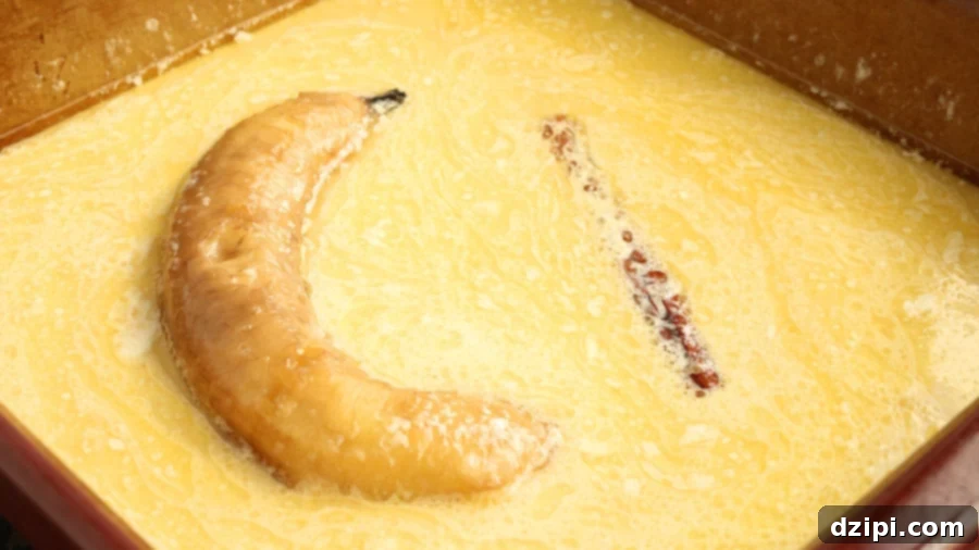 A peeled banana and a cinnamon stick are bathing in a baking dish filled with butter and cream.