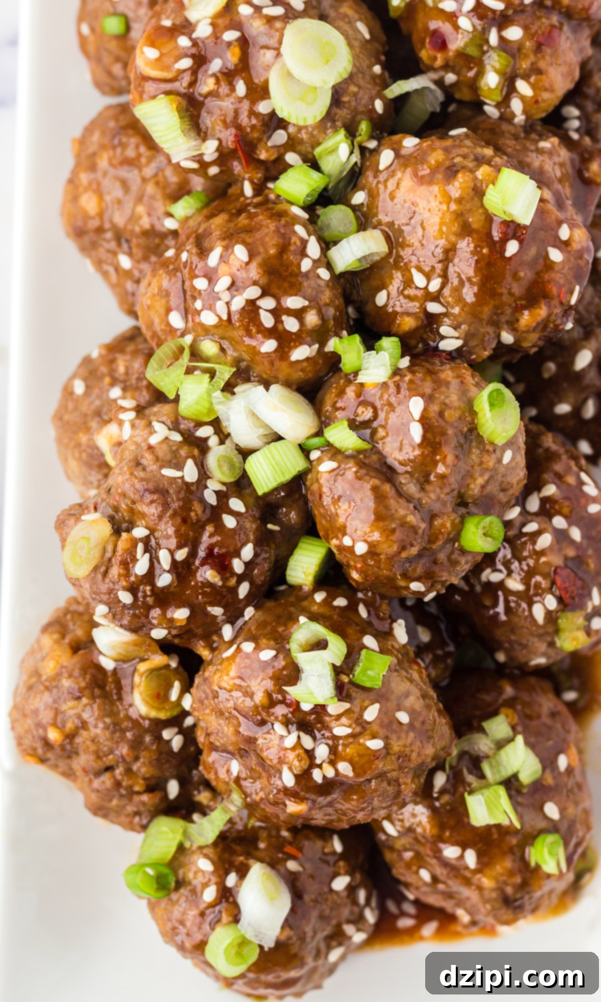 Overhead view of a few dozen Asian meatballs in brown sauce on a white plate. Garnished with sliced green onions and sesame seeds.