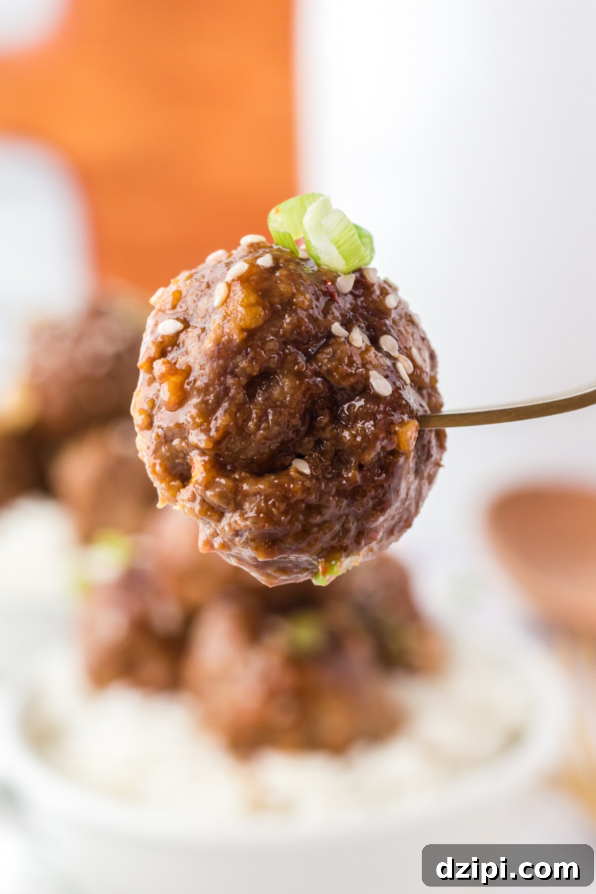 A fork holds up a single Asian meatball that's topped with sliced green onion and sesame seeds.