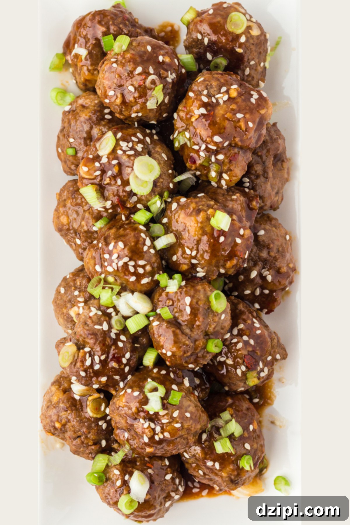 A white rectangular serving platter filled with saucy Asian meatballs garnished with green onions and sesame seeds.