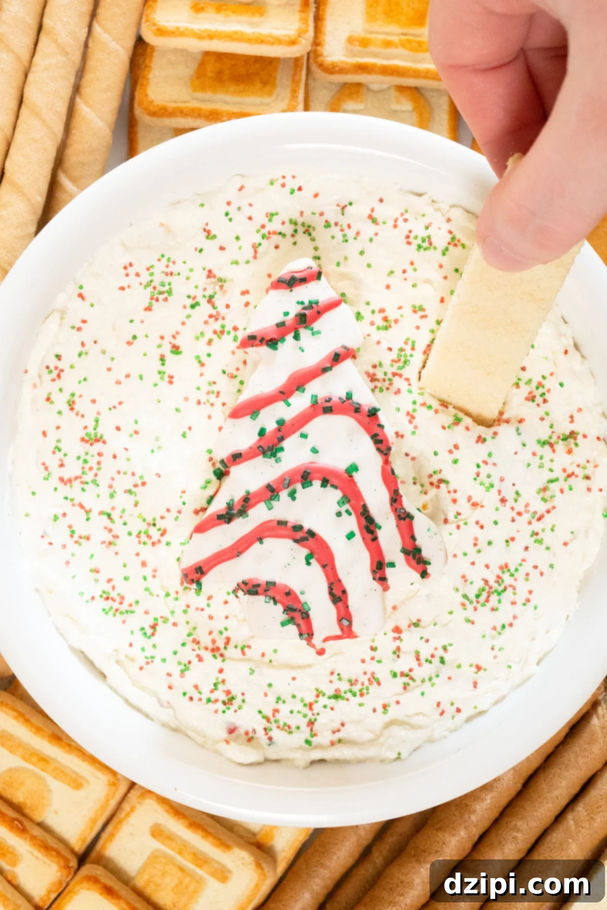 Holiday Tree Cake Dip 6 A hand gently dips a crispy vanilla sugar wafer into a bowl of the luscious Christmas Tree Cake Dip, showcasing its perfect creamy consistency.