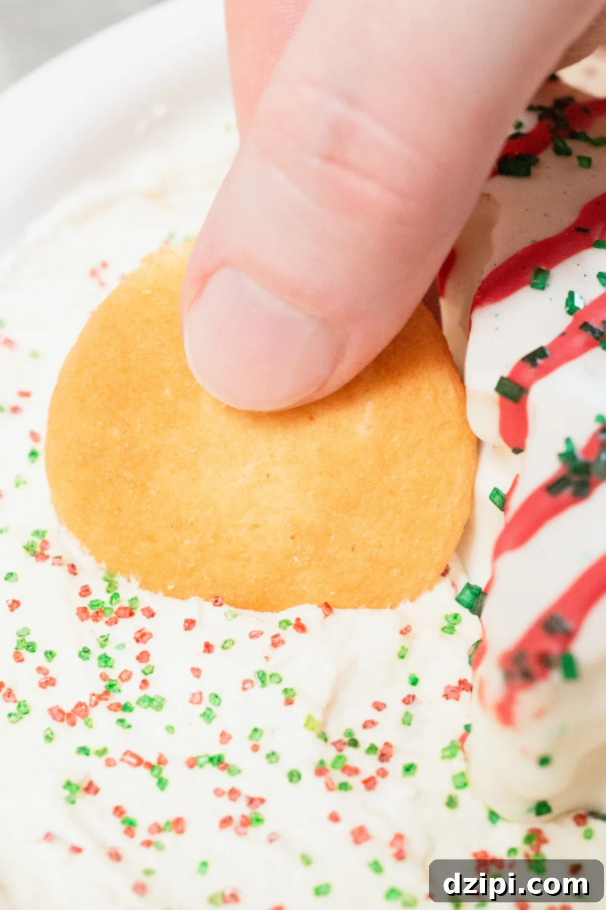 Holiday Tree Cake Dip 7 A close-up shot of a finger gracefully dipping a Nilla wafer into the creamy, inviting texture of Little Debbie's Christmas Tree Cake Dip.