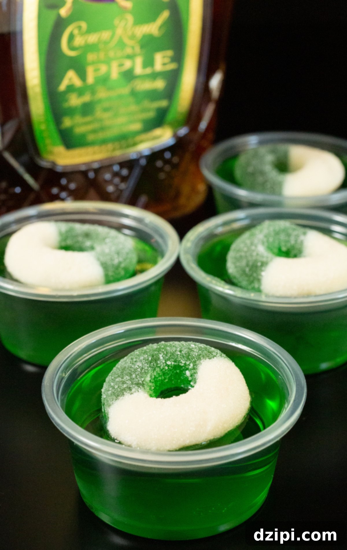Four 2oz plastic containers filled with green crown apple jello shots topped with a green apple candy ring.