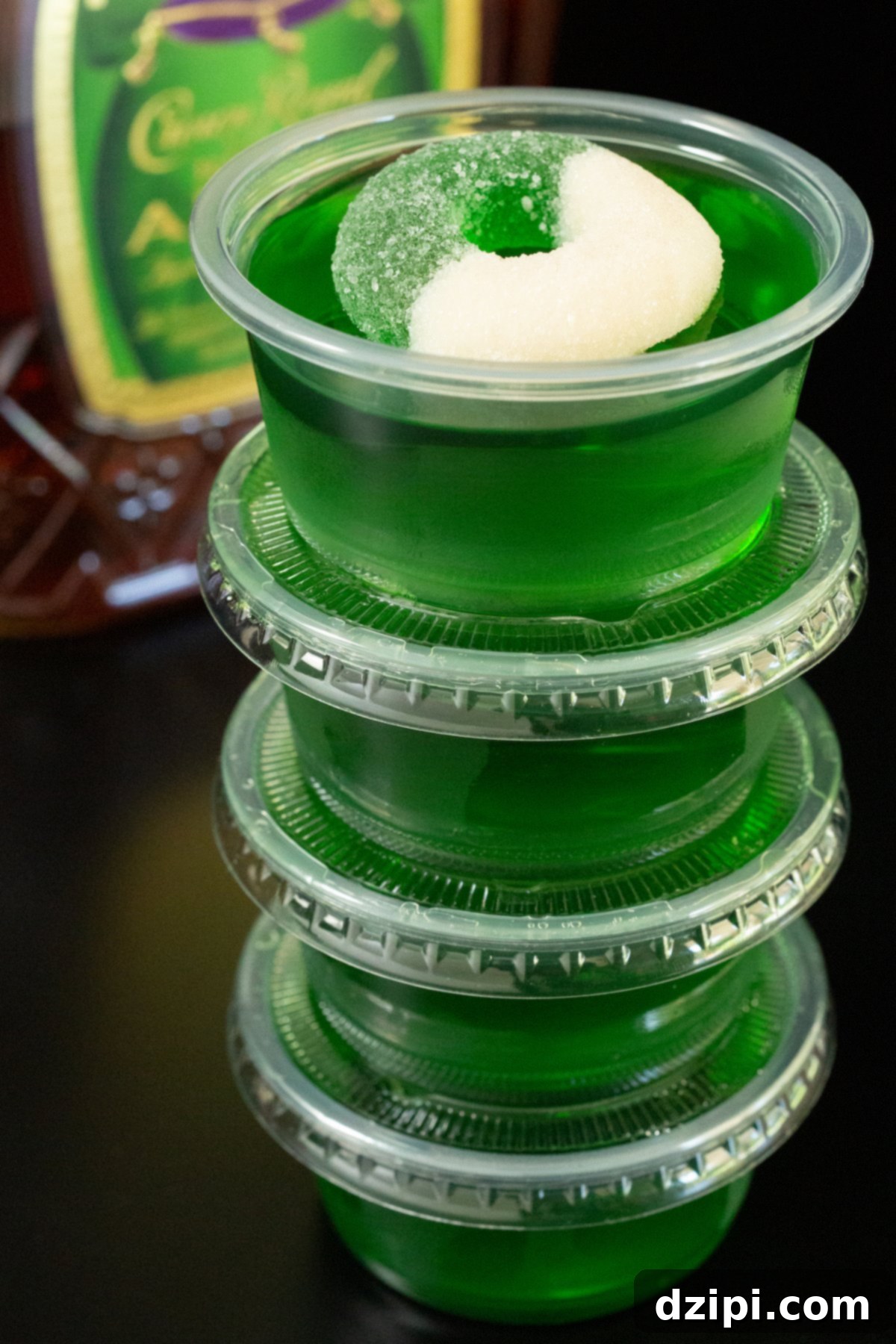 Four crown apple green jello shots stacked on top of each other. A bottle of Crown Apple is out of focus in the background.