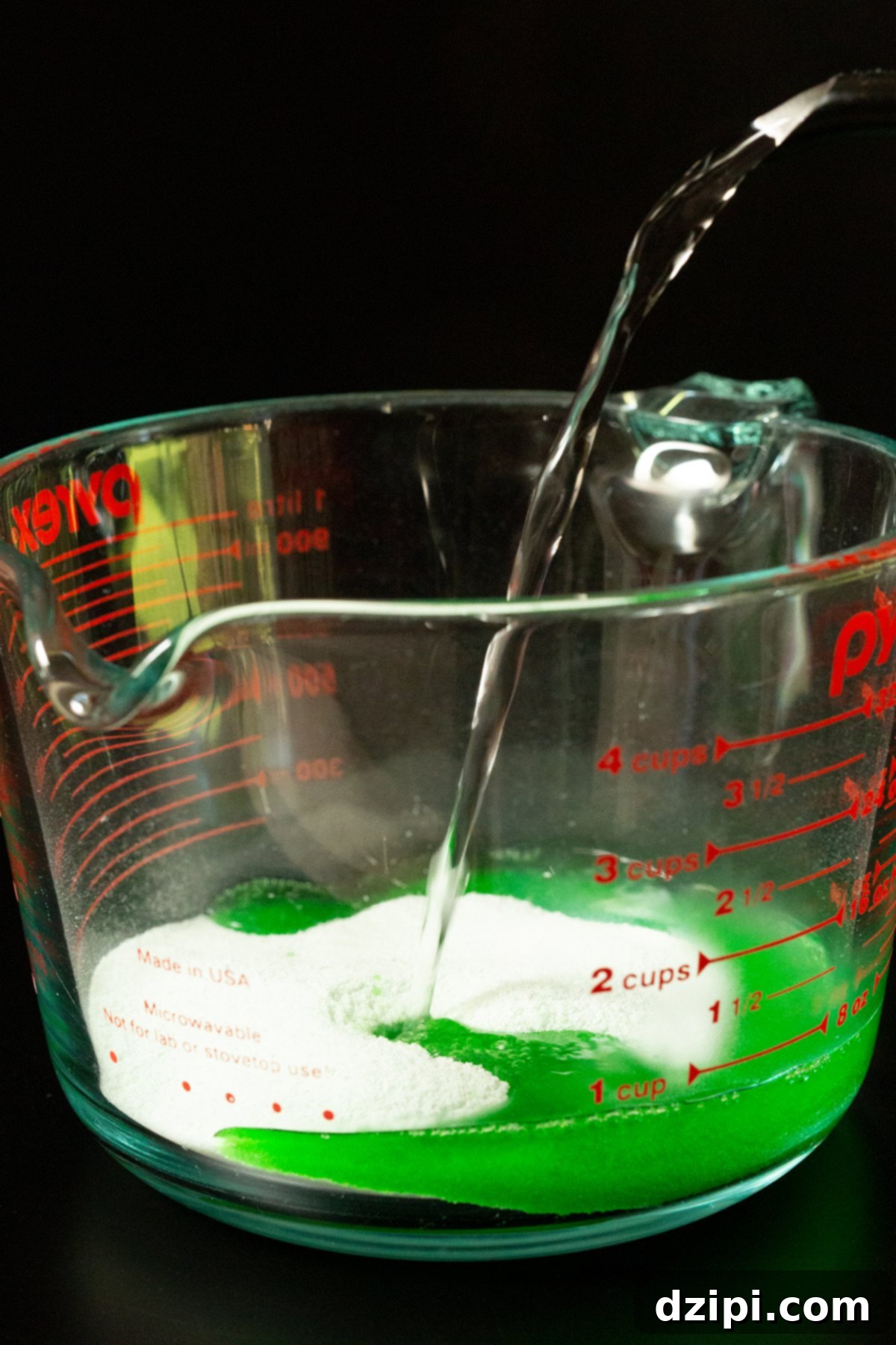 Hot water from a kettle being poured into a 4-cup glass measuring cup that has green apple gelatin powder in it.