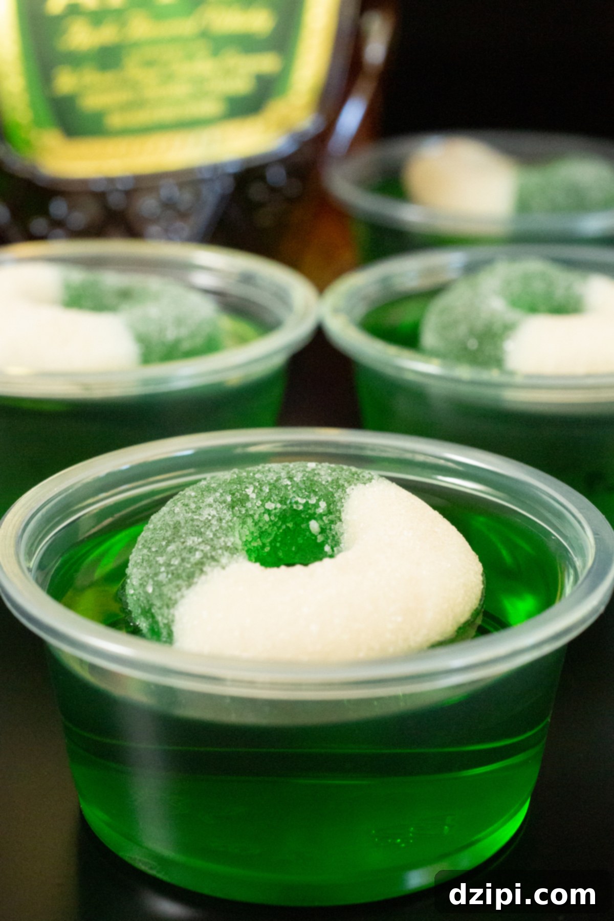 Close up of a 2oz plastic container filled with bright green Crown Apple jello shot that is topped with an apple candy ring.