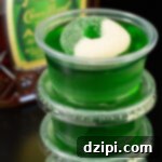 Four crown apple green jello shots stacked on top of each other. A bottle of Crown Apple is out of focus in the background.