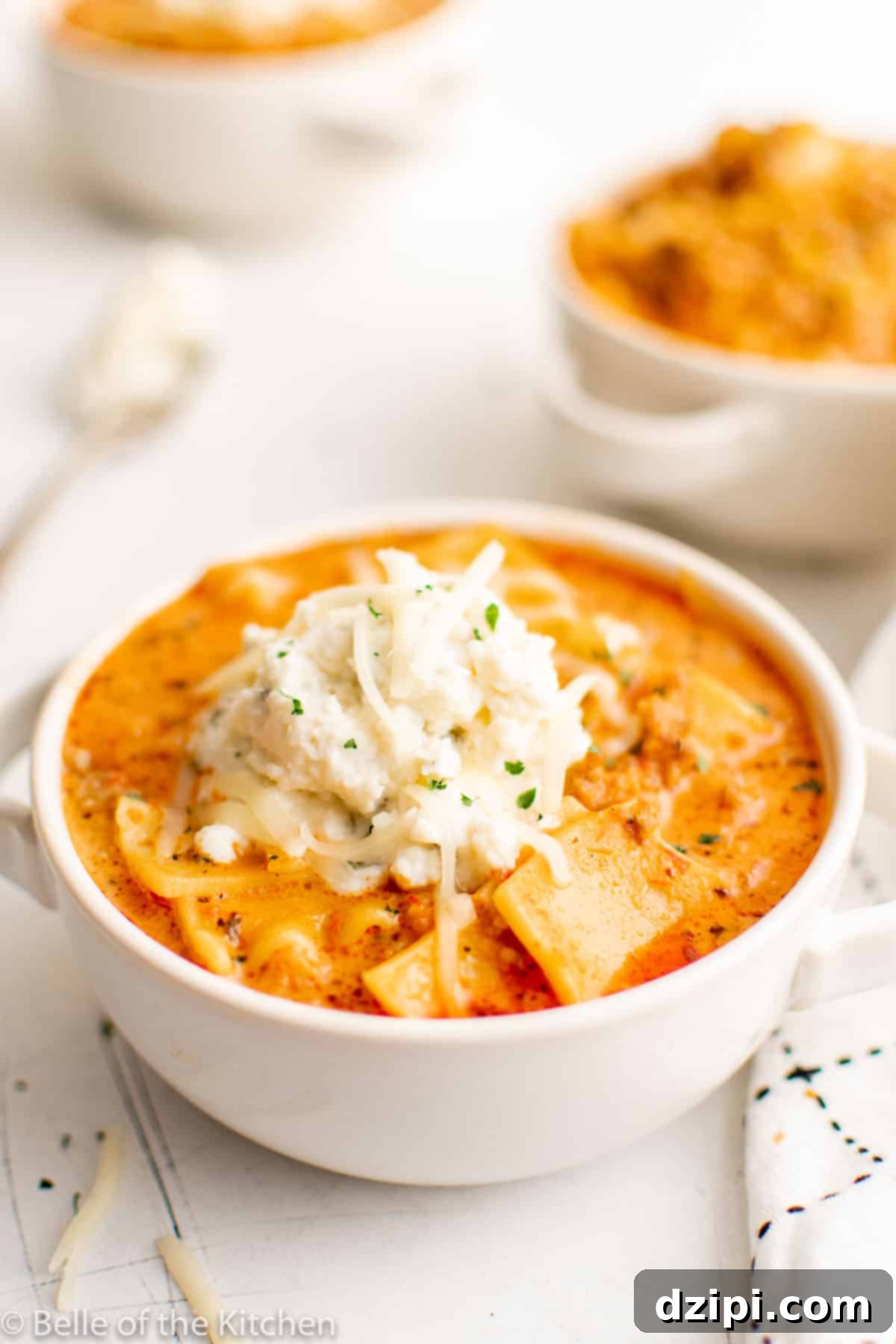 A pristine white bowl filled with savory lasagna soup, beautifully garnished with a creamy dollop of ricotta and a generous sprinkle of shredded cheese, promising a delightful meal.