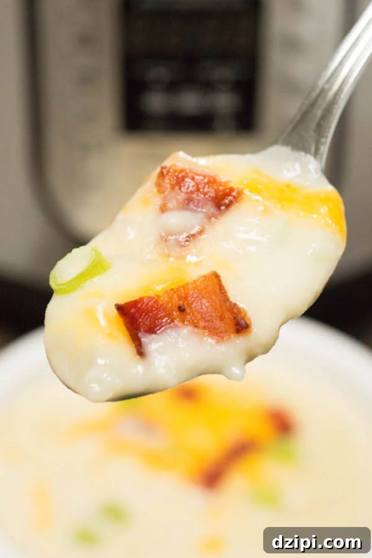 A spoonful of creamy Instant Pot loaded baked potato soup is perfectly in focus, with the pressure cooker and a full bowl of the hearty soup softly blurred in the background, highlighting its delicious texture.