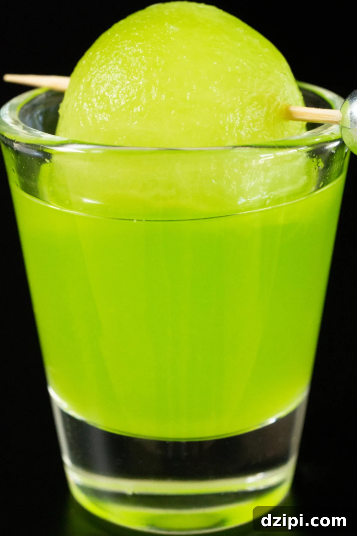 A single shot glass filled with a bright green melon ball shot, garnished with a honeydew melon ball on a toothpick.
