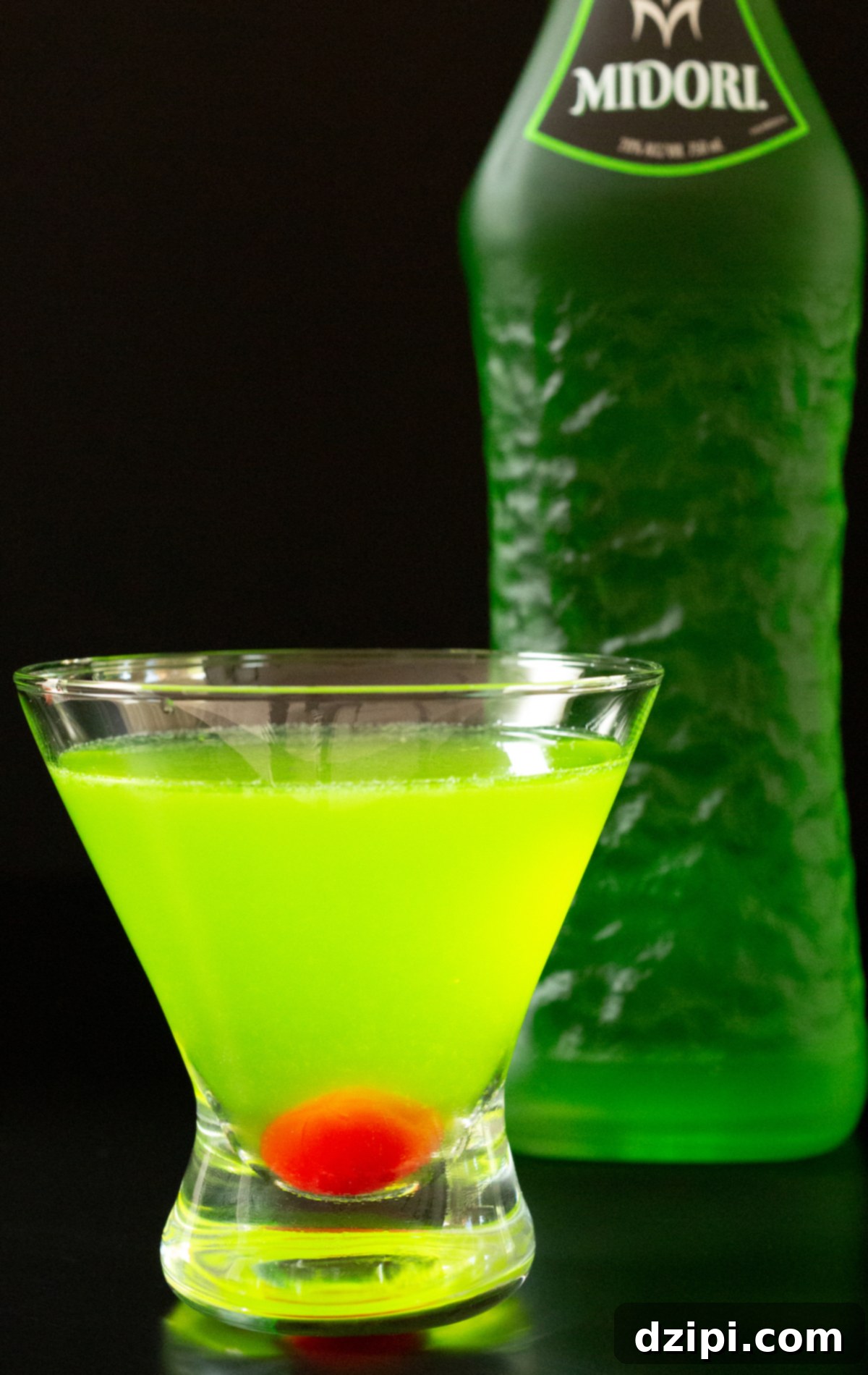 A stemless martini glass filled with a neon green Japanese slipper cocktails that has a maraschino cherry in the bottom. A bottle of Midori is out of focus in the background.