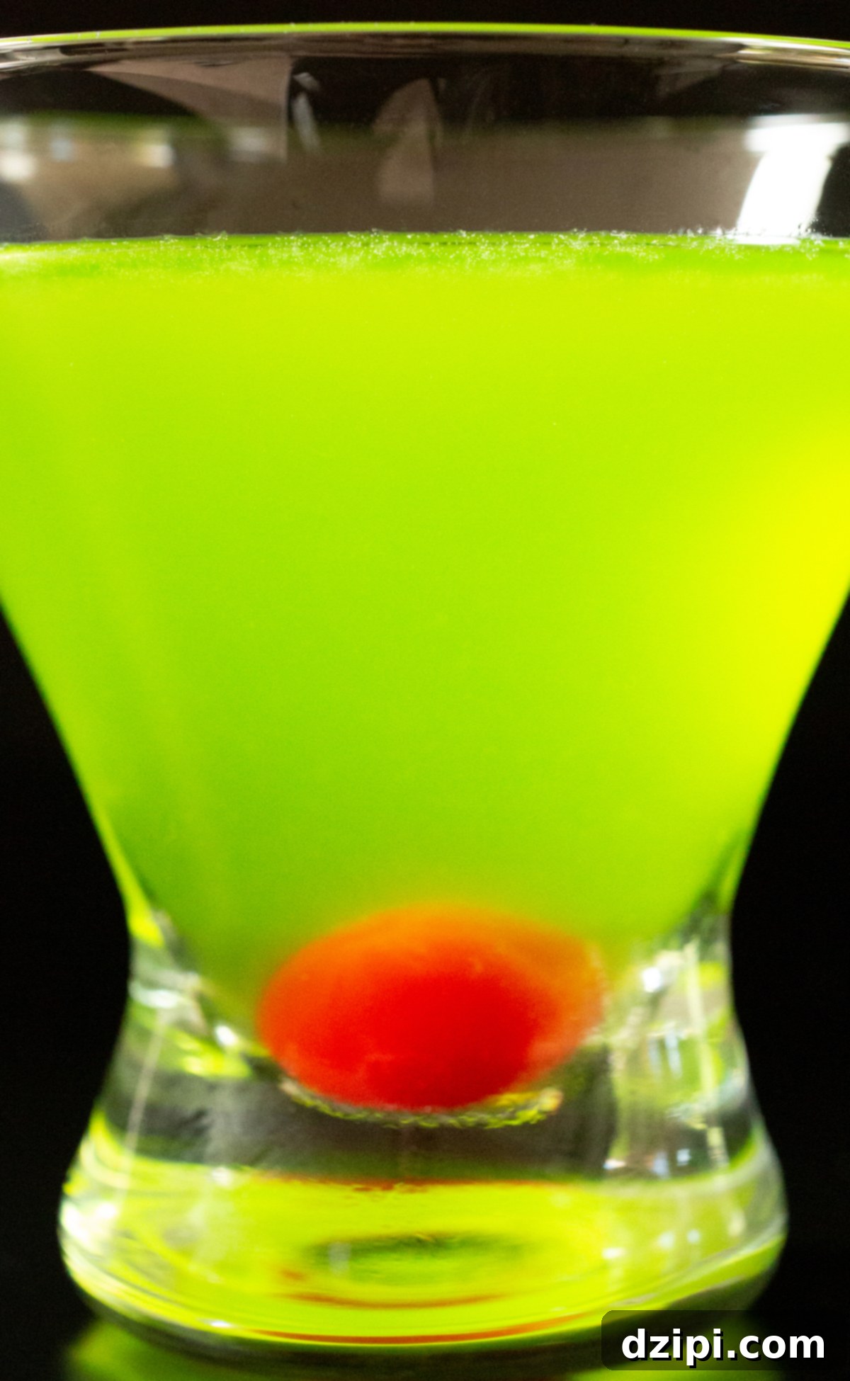 A stemless martini glass is filled with a neon green japanese slipper cocktail. There is a stemless maraschino cherry in the bottom of the glass.