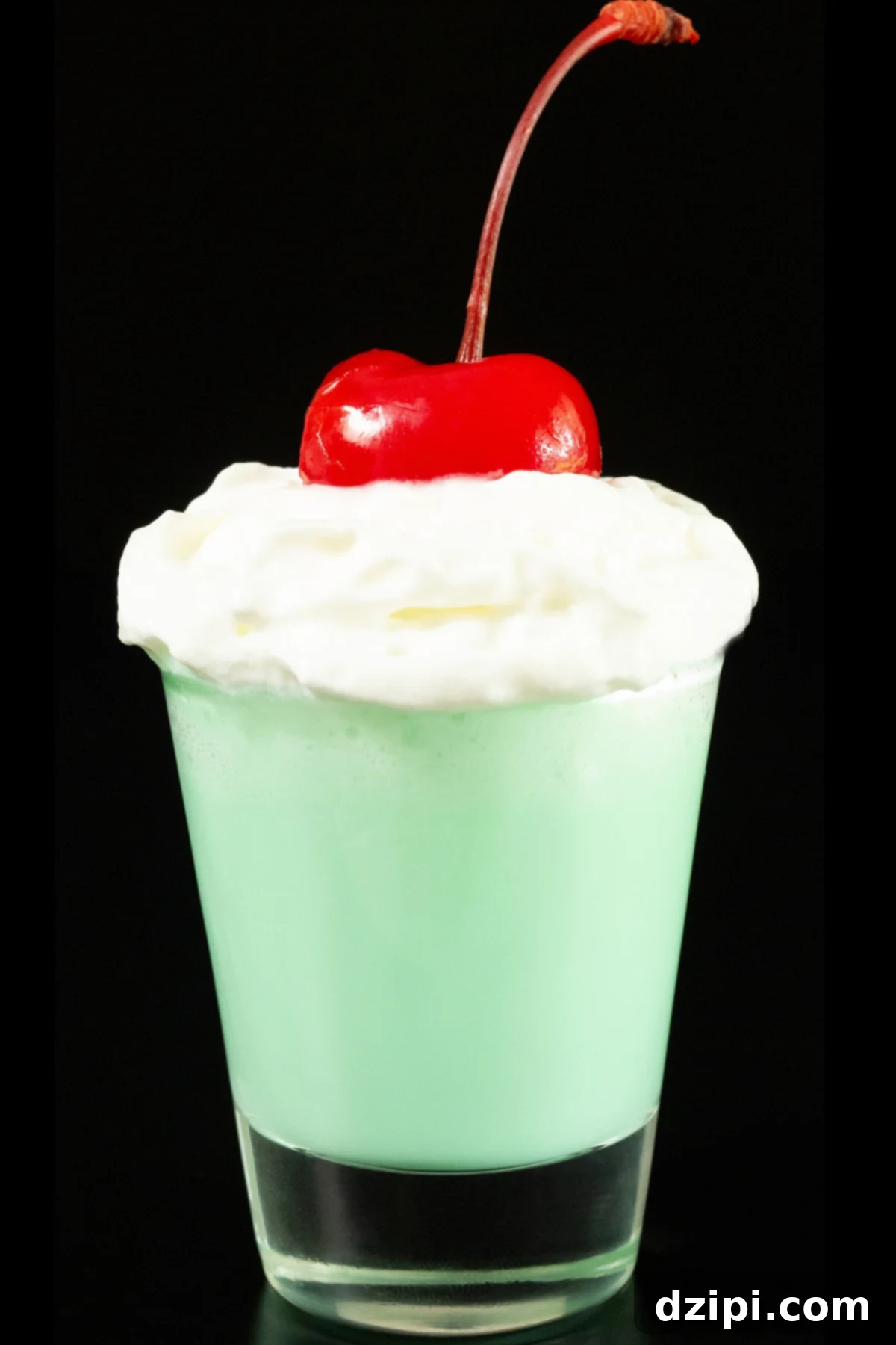 Emerald Elixir 3 A single shot glass on a white background is filled with mint colored shamrock shot mixture that's topped with whipped cream and a maraschino cherry, representing the perfect St. Patrick's Day drink.