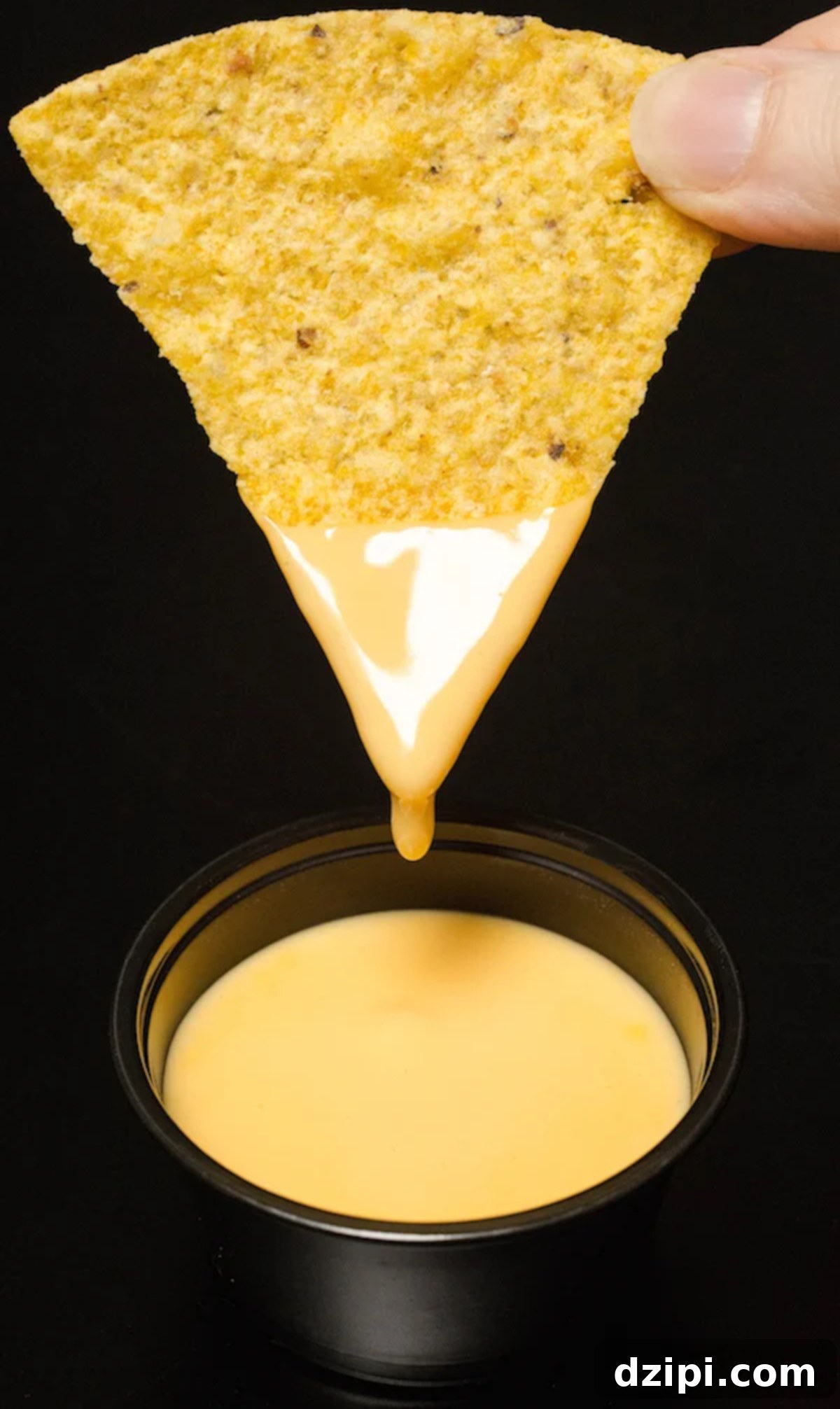 A hand holding a single, crispy tortilla chip, perfectly coated in a generous dip of creamy nacho cheese sauce.