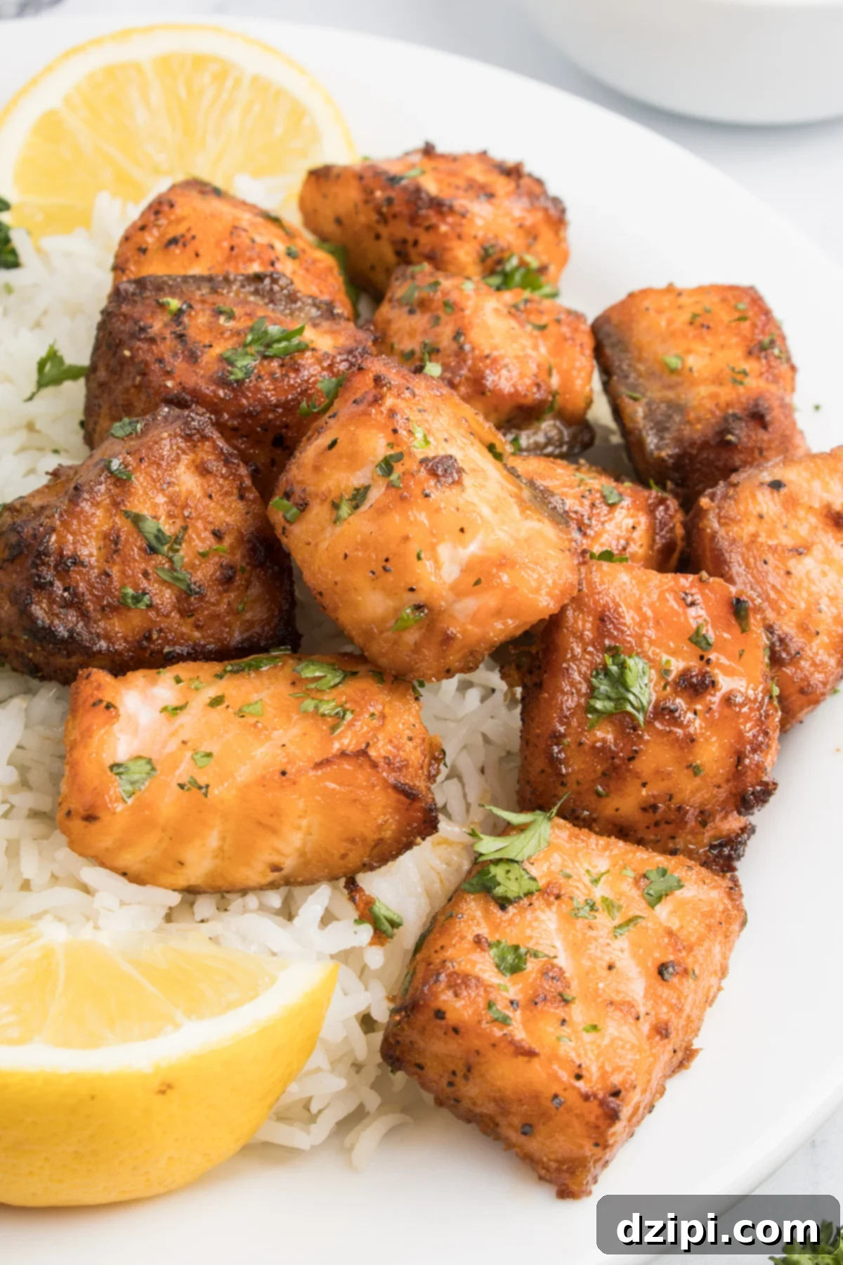 Bite sized pieces of salmon cooked in the air fryer on a white dinner plate with rice and lemon slices. 