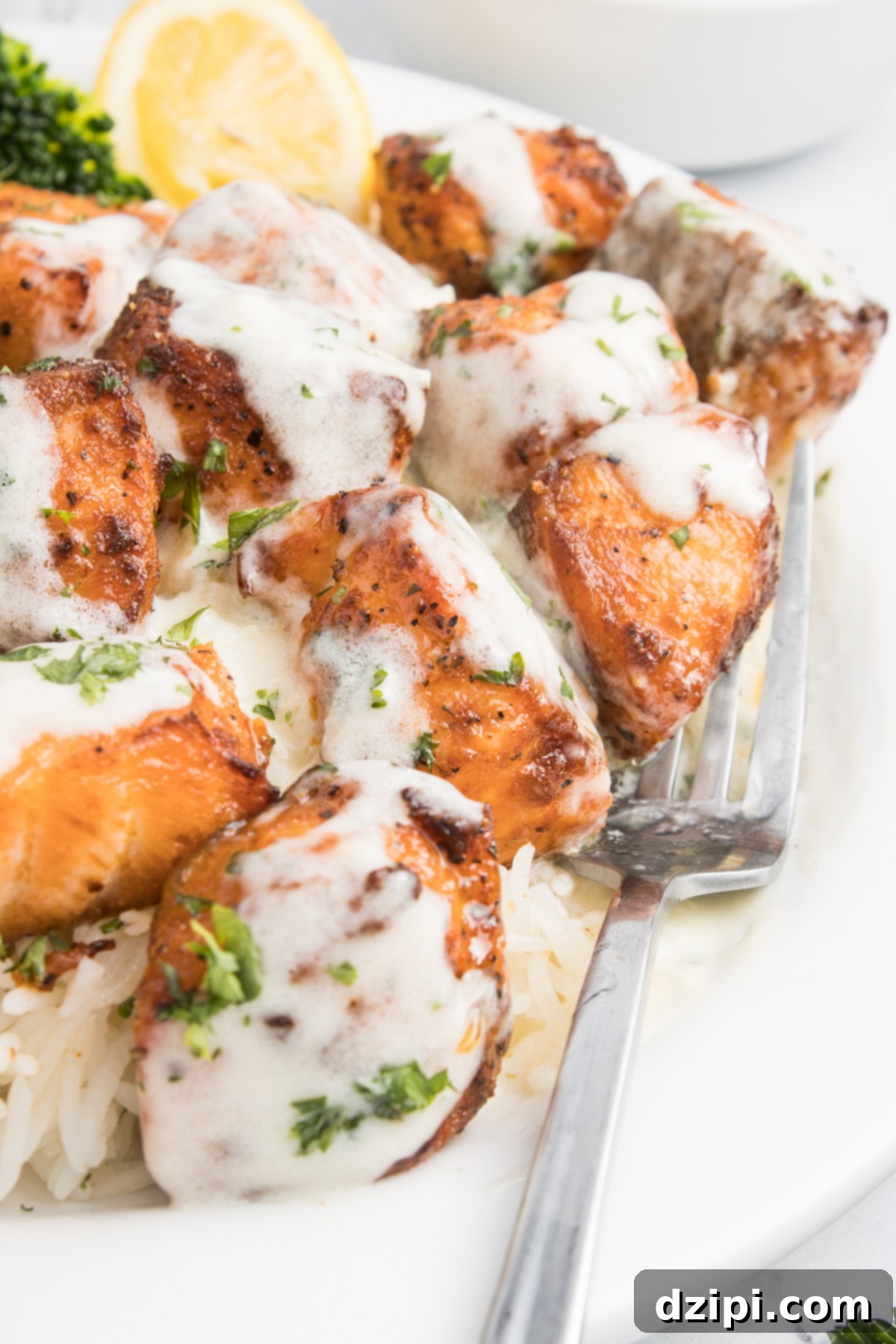 Close up of garlic salmon bites covered in garlic cream sauce next to a fork. A piece of broccoli and lemon slice are out of focus in the background.