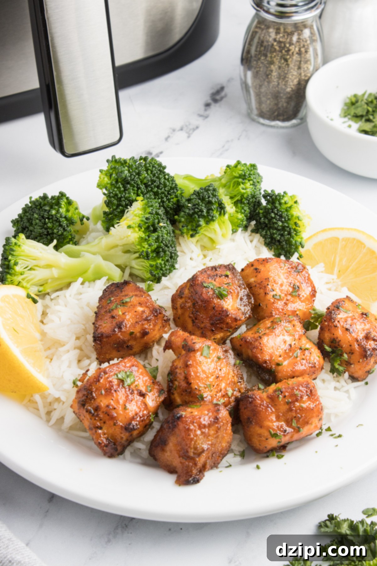 A white dinner plate with salmon bites, white rice, broccoli, and lemon slices in front of an air fryer.