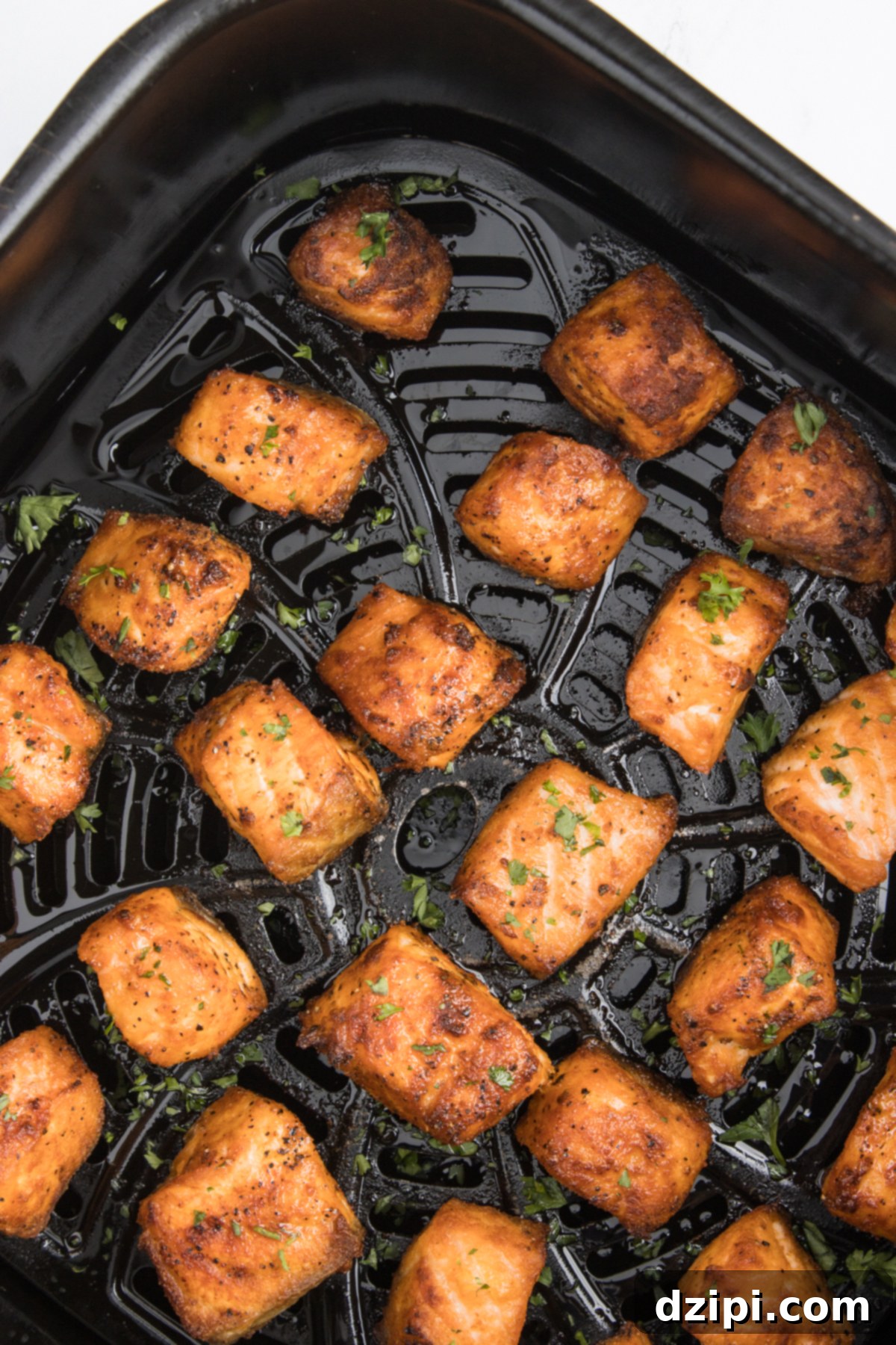 An air fryer basket filled with cooked salmon bites that are garnished with fresh chopped parsley.