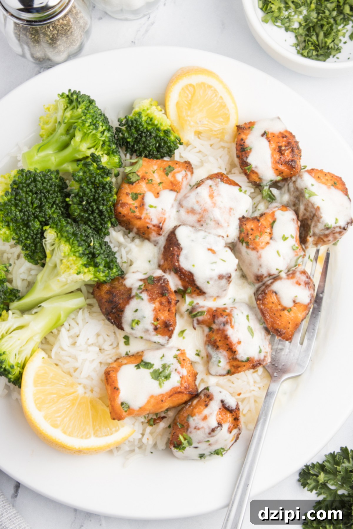 A white dinner plate with salmon bites covered in garlic cream sauce with white rice and broccoli. There are also two lemon slices, one has been squeezed.