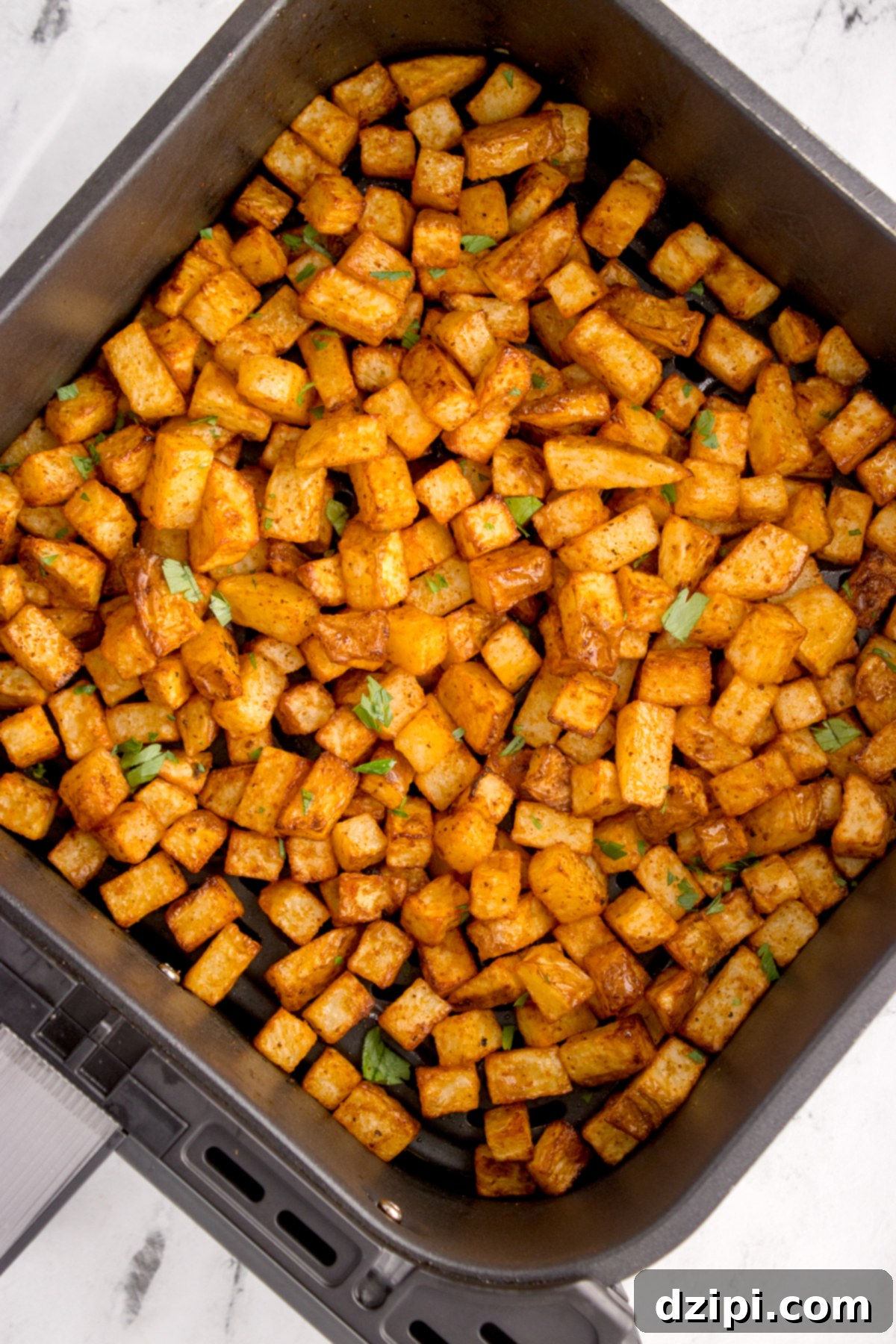 An air fryer basket filled with cooked diced breakfast potatoes that are garnished with chopped parsley.