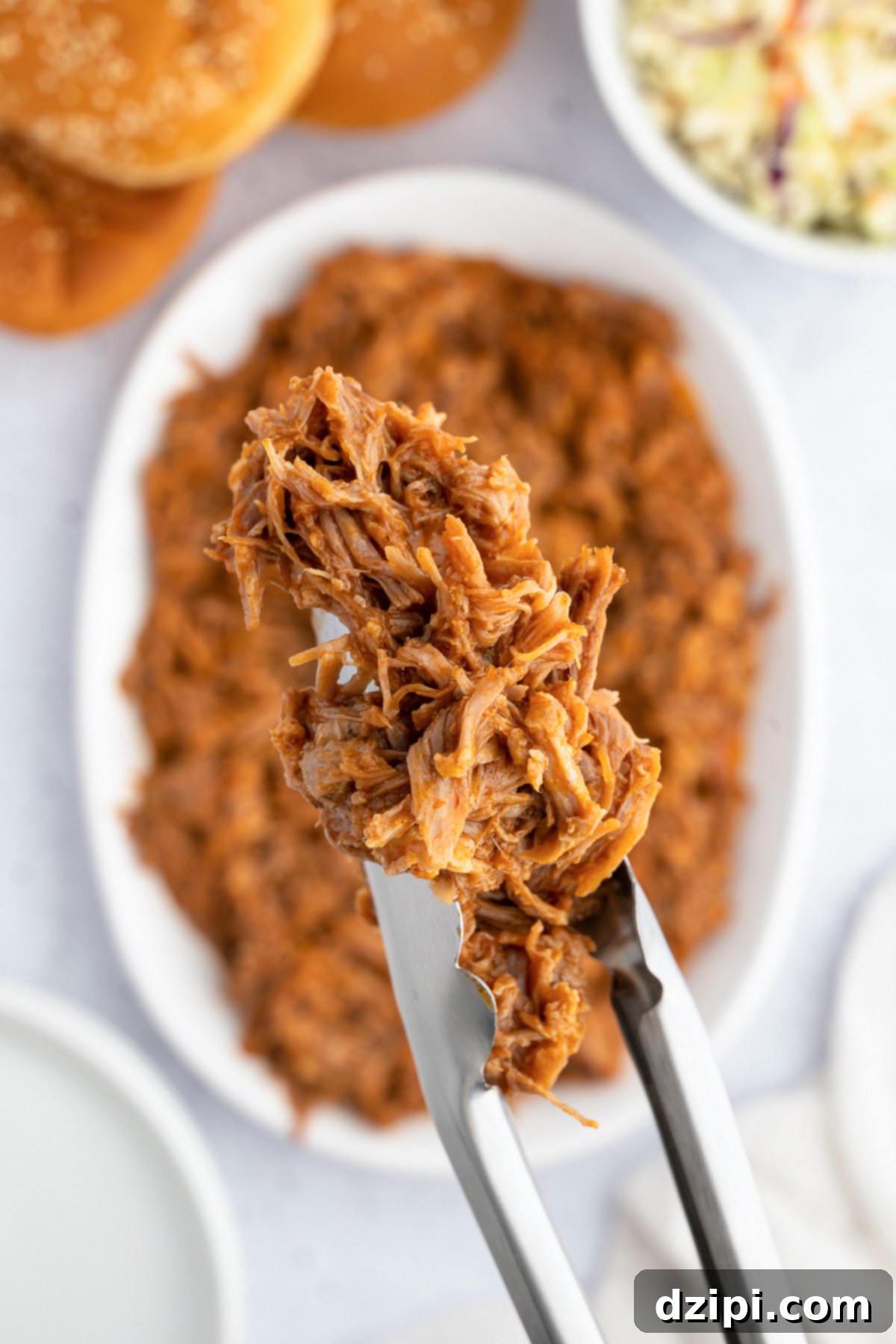 Juicy Instant Pot Pulled Pork 3 Serving tongs lifting a generous portion of freshly shredded Instant Pot pulled pork from a full dish, with the focus on its moist, tender texture.