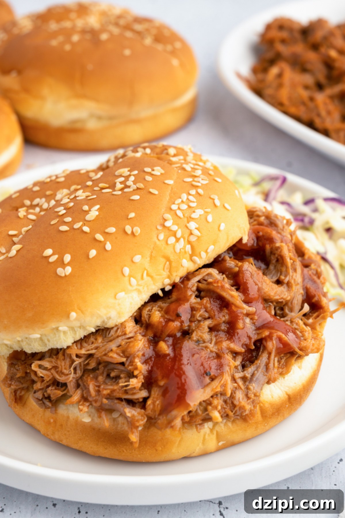 Juicy Instant Pot Pulled Pork 7 A perfectly assembled pulled pork sandwich takes center stage in the foreground, with blurred hamburger buns and more pulled pork waiting in the background.