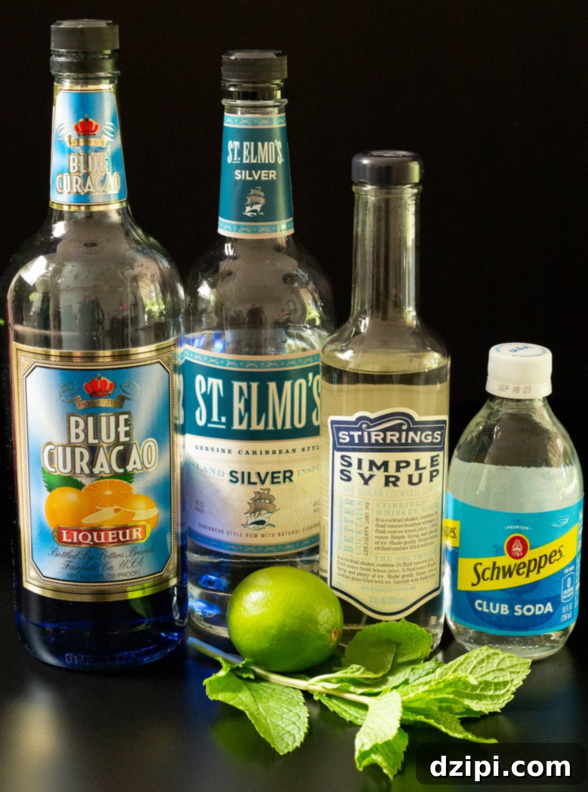 Sapphire Sip 4 A collection of Blue Mojito ingredients laid out on a dark surface: a bottle of clear white rum, blue curacao, simple syrup, a fresh lime, a bundle of mint leaves, and a bottle of club soda.