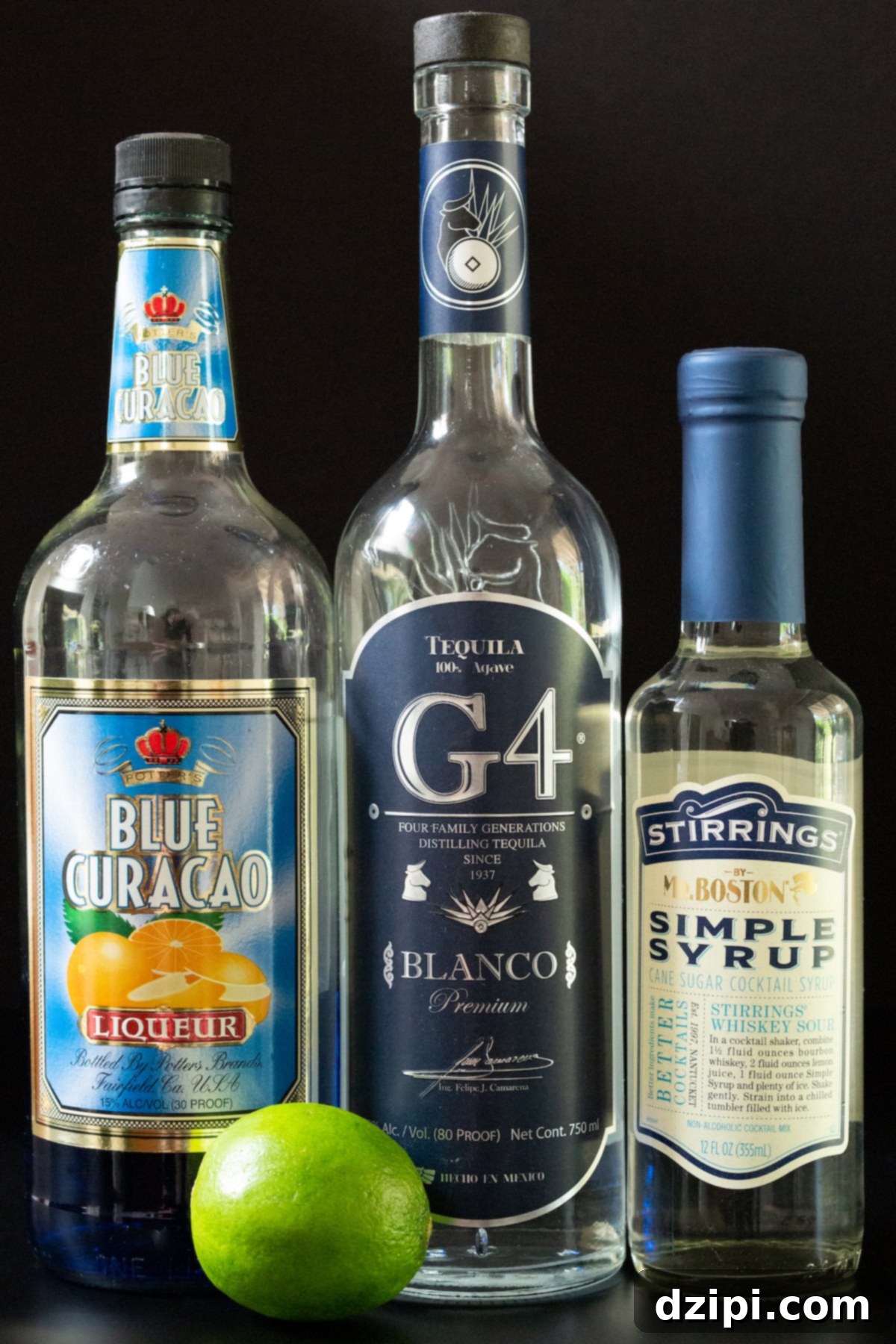 Ingredients for a Blue Margarita laid out on a table: tequila, lime juice, blue curacao, and simple syrup.