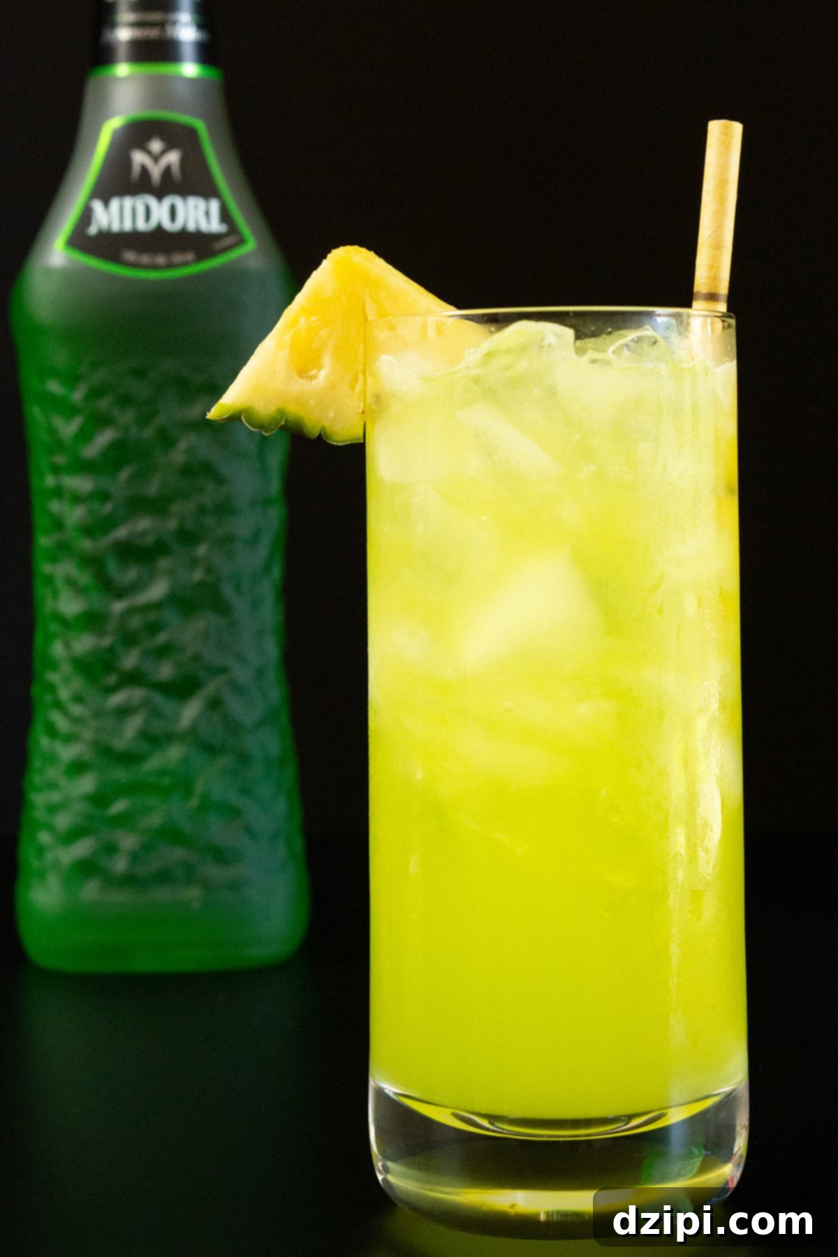 A highball glass filled with a bright yellow-green June Bug cocktail, garnished with a fresh pineapple triangle. In the soft-focus background, a bottle of Midori melon liqueur hints at the drink's key ingredient, creating an inviting tropical atmosphere.