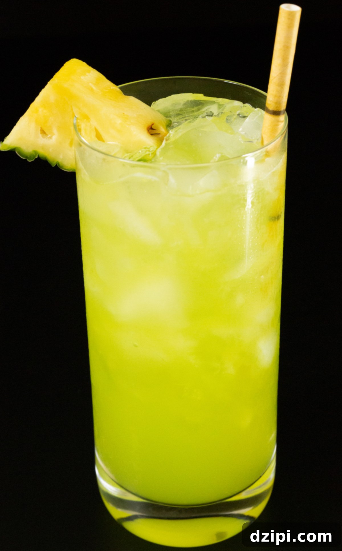 A highball glass showcasing a perfectly crafted yellow-green June Bug cocktail. It features a charming bamboo-style paper straw and a fresh pineapple triangle elegantly attached to the left rim as a vibrant garnish, inviting a taste of the tropics.