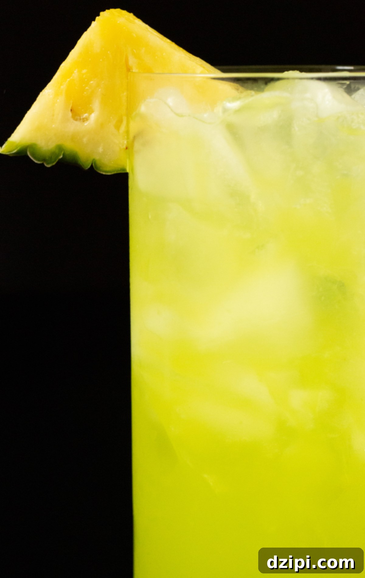 A highball glass filled with a vibrant yellow-green June Bug cocktail, elegantly garnished with a fresh pineapple triangle. The refreshing appearance invites a tropical tasting experience.