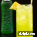 A highball glass filled with a bright yellow-green cocktail that's garnished with a pineapple triangle. A bottle of Midori is out of focus in the background.