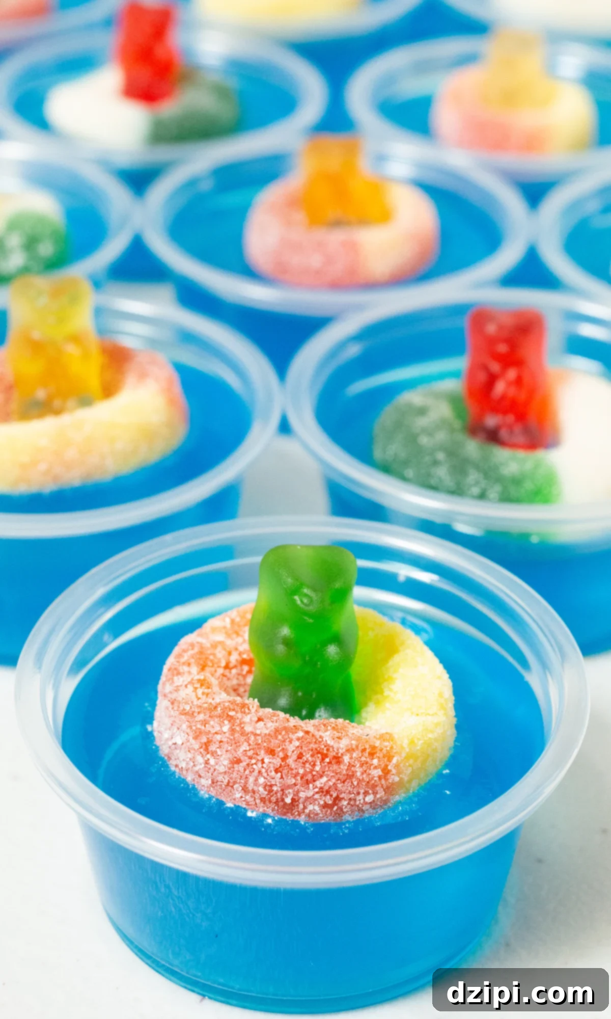 Nine 2oz plastic containers filled with bright blue jello, each topped with a colorful gummy bear nestled in a gummy ring, ready for a pool party.