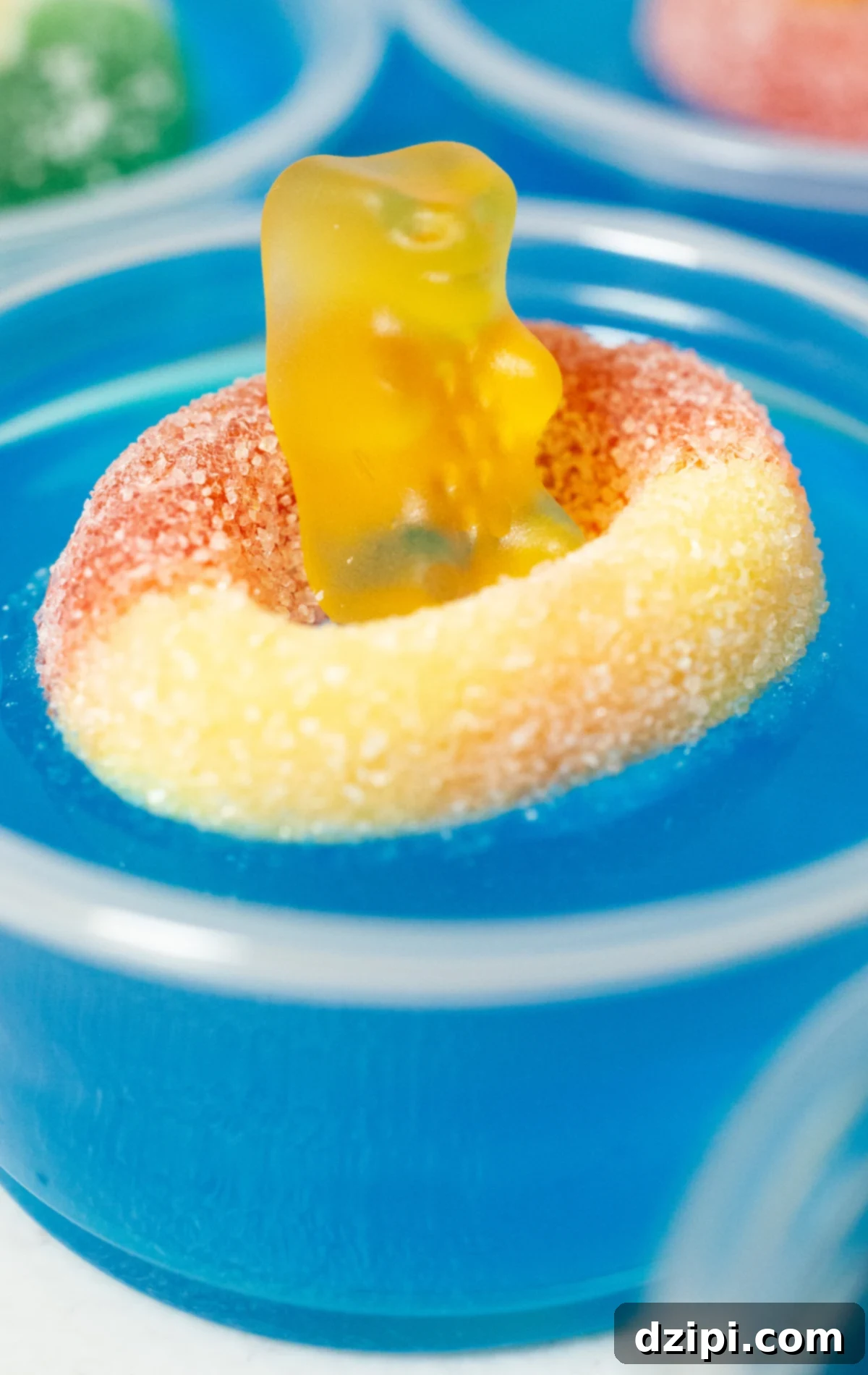 A single pool party jello shot featuring bright blue gelatin, topped with a peach gummy ring and a yellow gummy bear, creating a charming miniature pool scene.