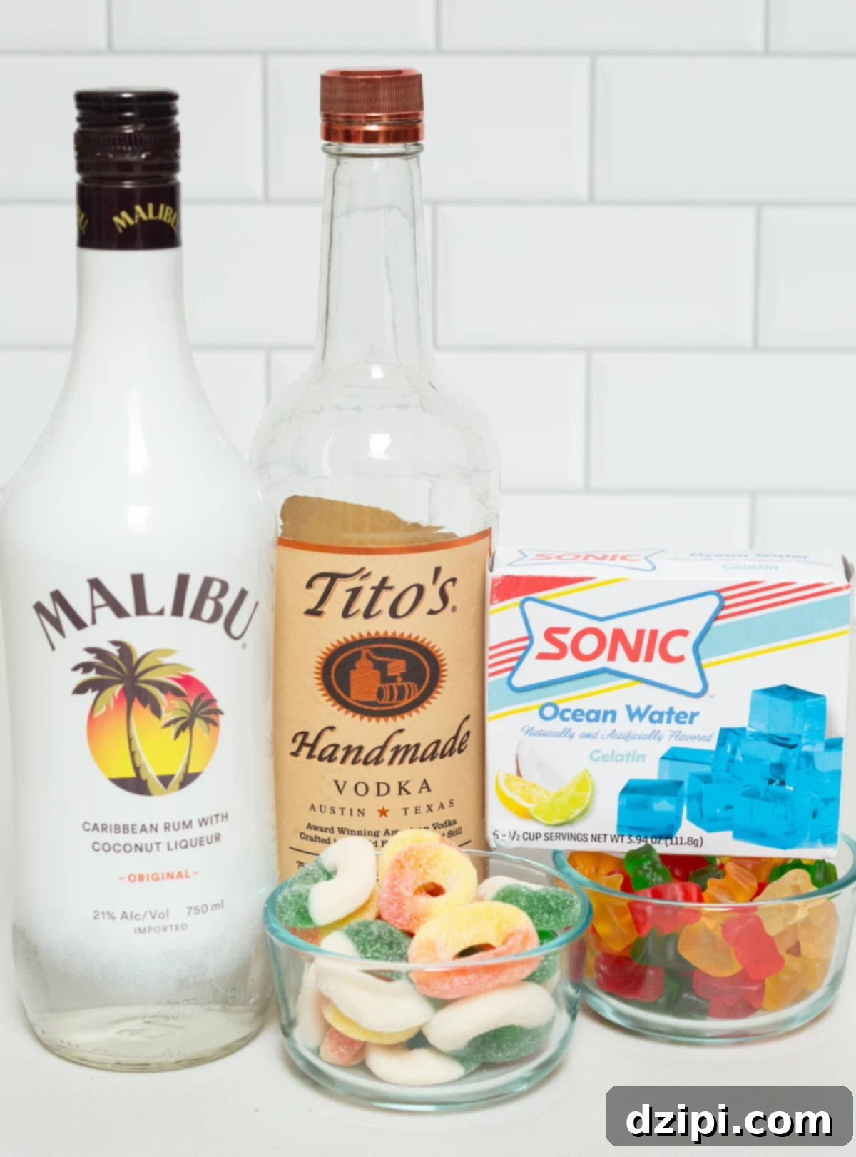 A collection of ingredients laid out on a white background, including bottles of Malibu rum and vodka, blue gelatin mix, various gummy rings, and an assortment of gummy bears, all necessary for making pool party jello shots.