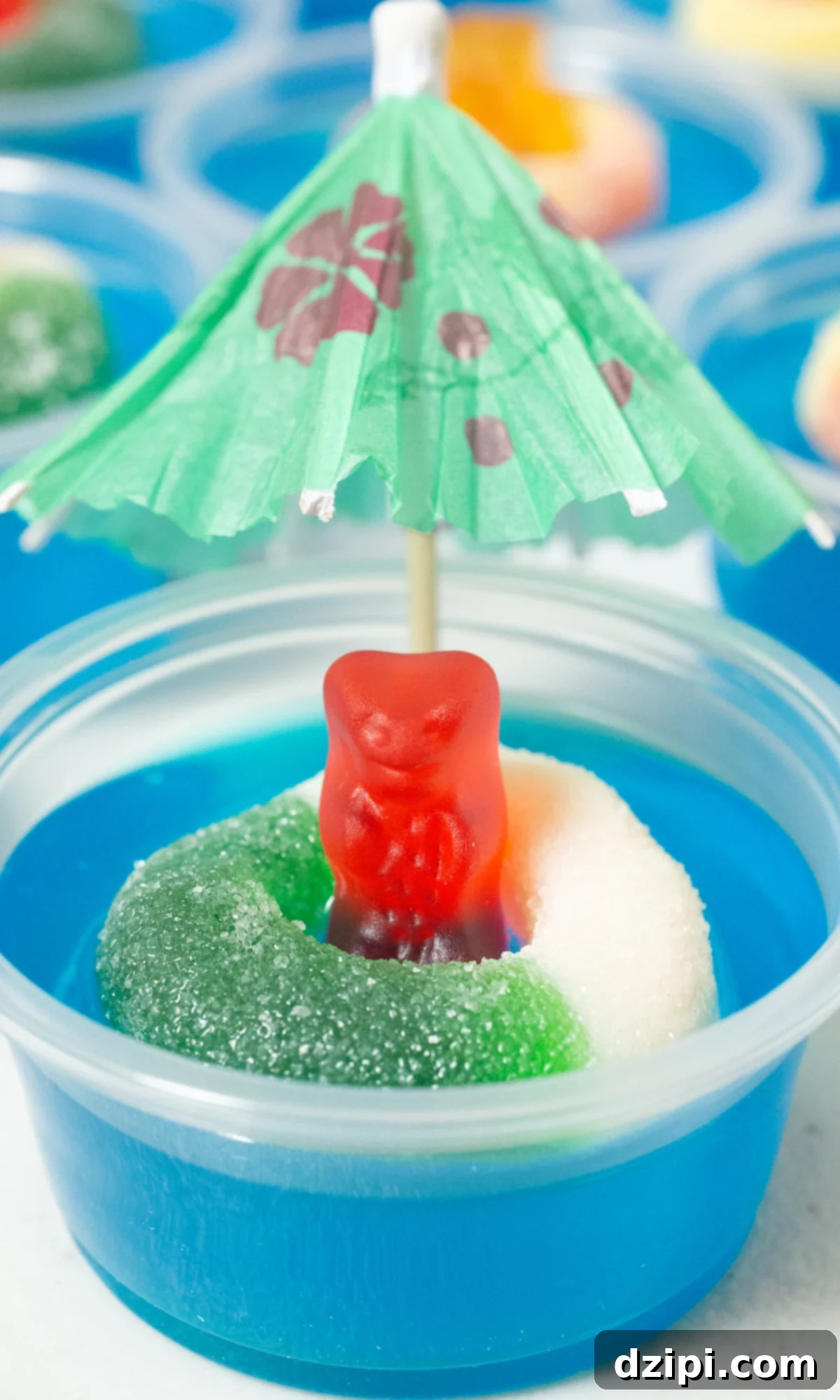 A single 2 oz plastic container filled with a blue jello shot, garnished with a gummy bear happily floating inside a gummy ring, and topped with a tiny cocktail umbrella for extra charm.