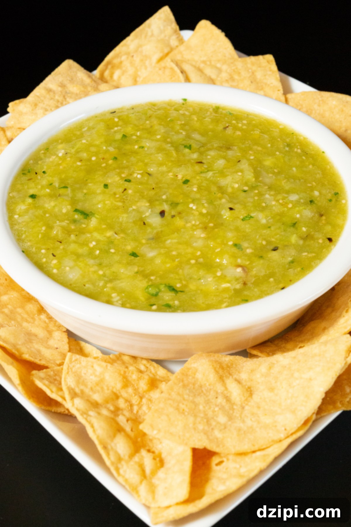 A white serving bowl brimming with vibrant green Hatch chile salsa, artfully arranged on a rustic serving plate, surrounded by golden tortilla chips ready for dipping. The scene evokes a fresh, delicious Mexican appetizer.