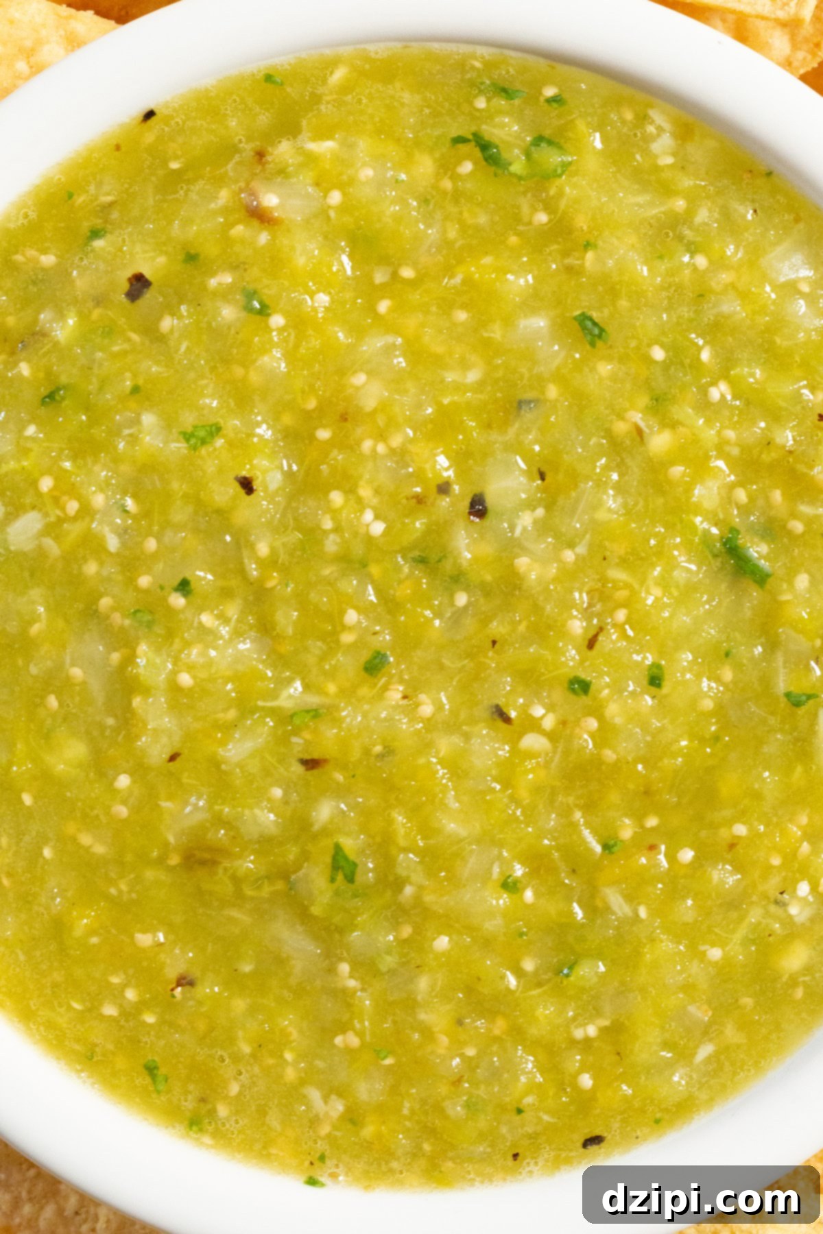 A close-up view of a white serving bowl filled with beautifully textured green Hatch chile salsa, highlighting its vibrant color and fresh ingredients. This spicy, tangy, and bright salsa is perfect as a dip or topping.