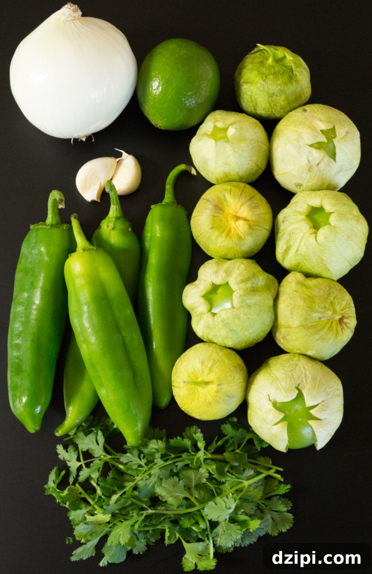 A vibrant display of fresh ingredients for green chile salsa laid out on a dark background: plump Hatch chiles, firm tomatillos, a quartered white onion, aromatic garlic cloves, and a bright green lime, with a bunch of fresh cilantro. All elements are ready for preparation.