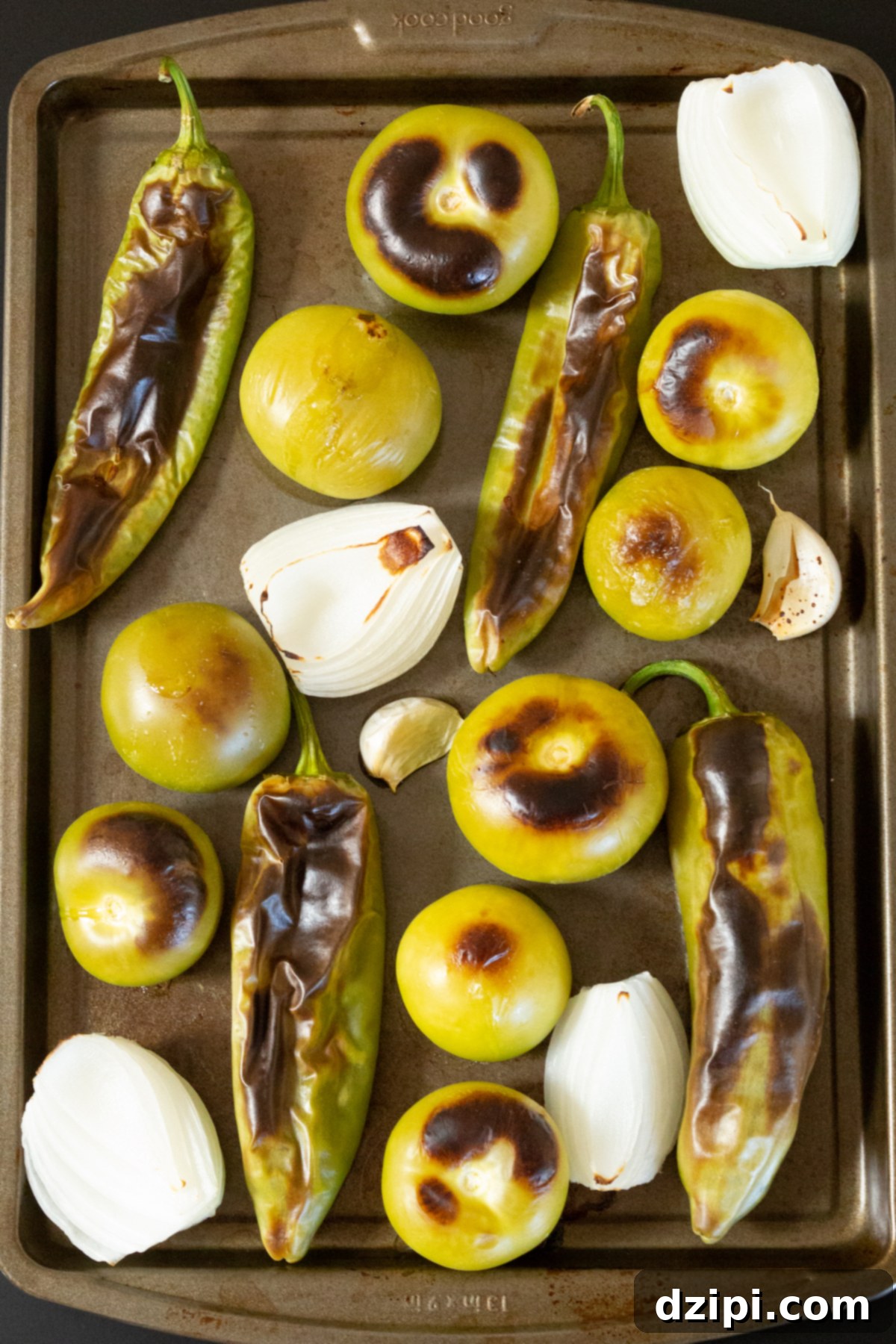 A baking sheet neatly arranged with freshly roasted Hatch chiles, plump tomatillos, quartered white onion, and cloves of garlic, all perfectly charred and softened from the broiler, ready for blending into a delicious salsa.