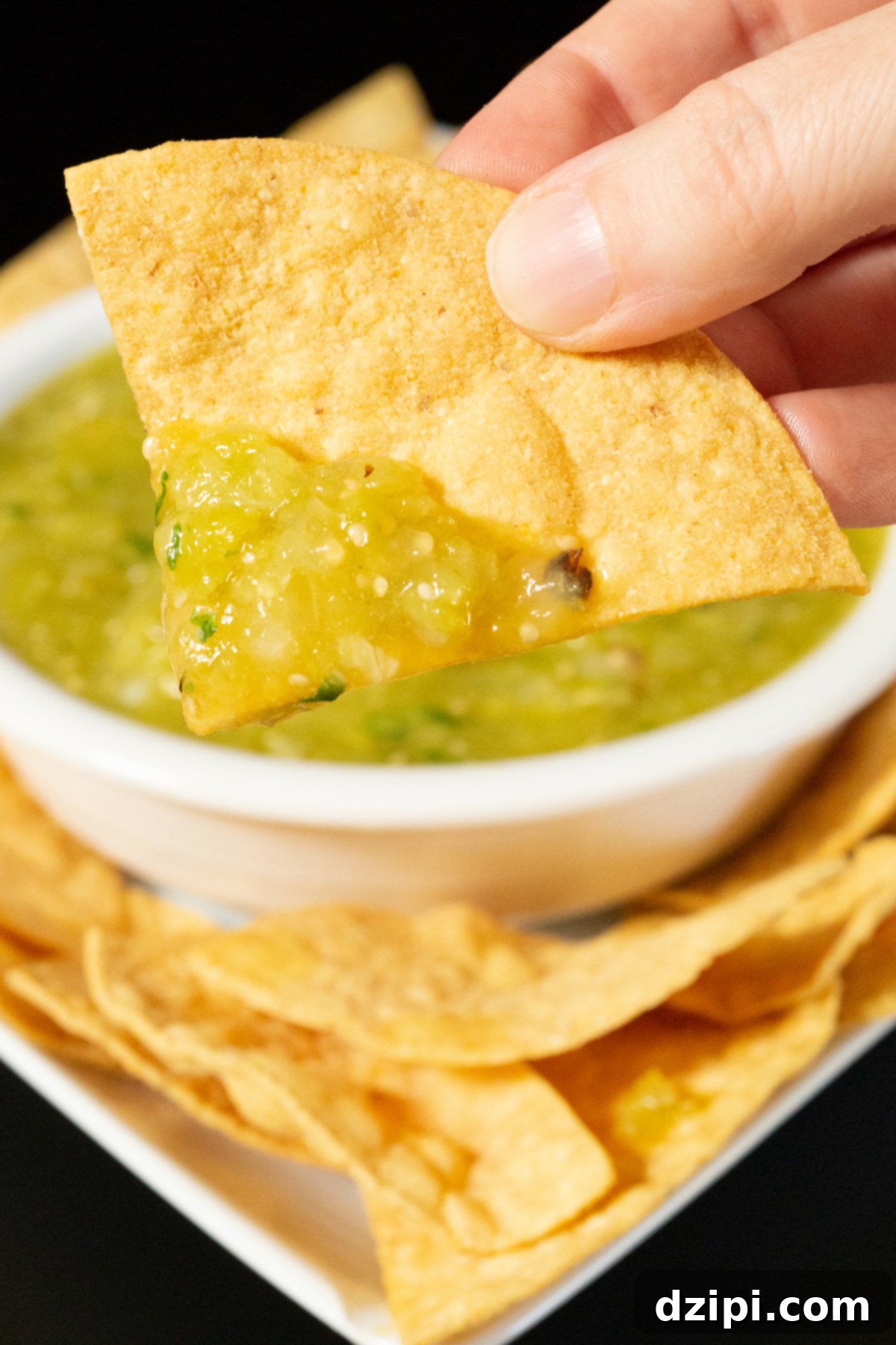 A hand holds up a single crispy tortilla chip, generously scooped with vibrant green Hatch chile salsa. In the softly blurred background, a bowl holds the remaining salsa, inviting more delicious dips. This image perfectly captures the joy of tasting freshly made salsa verde.