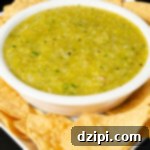A white serving bowl filled with green Hatch chile on a serving plate surrounded by tortilla chips.