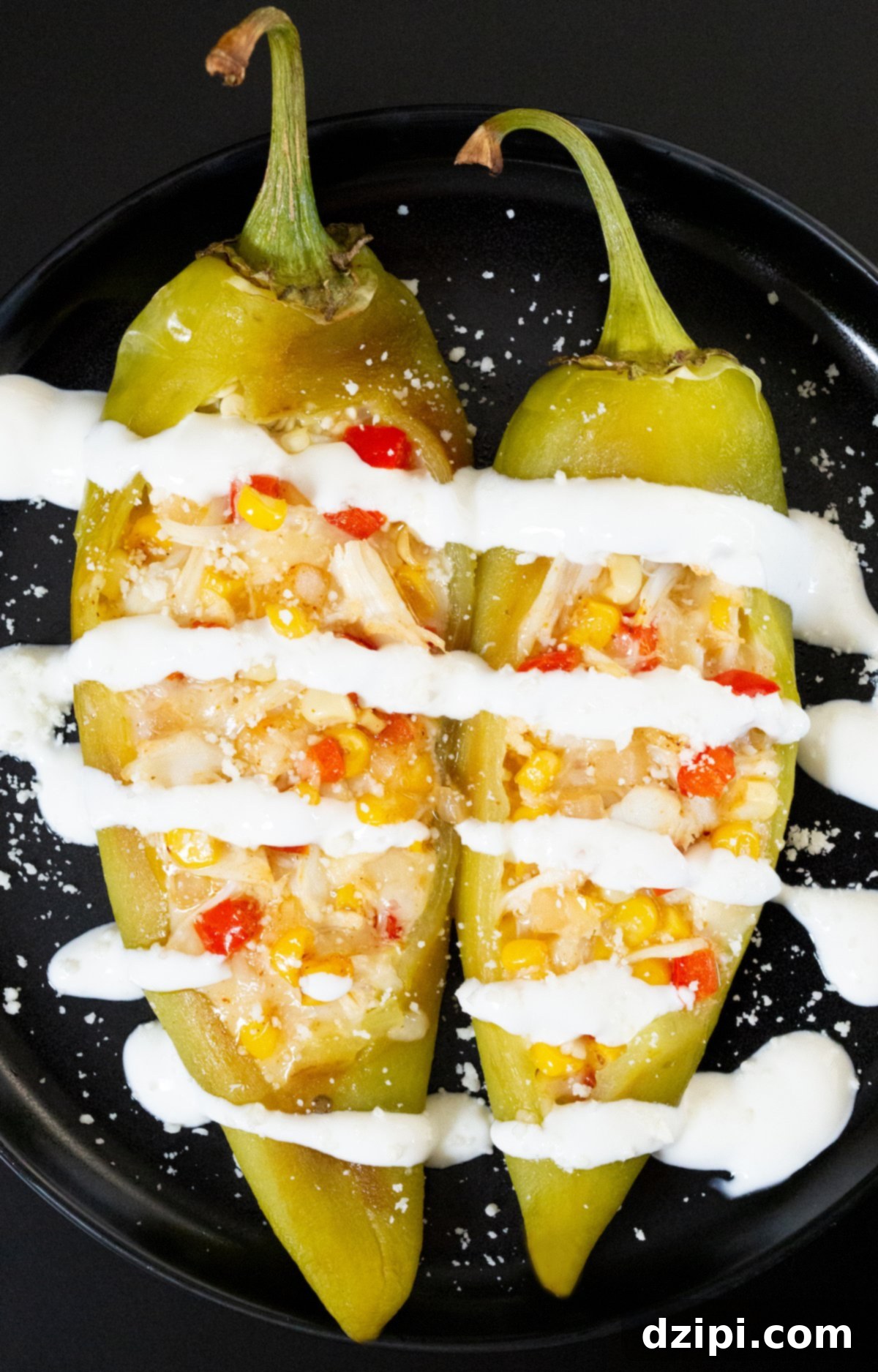 A small black plate with two large hatch peppers that have been stuffed with crab, corn, red bell pepper, and cheese. They ache been drizzled with lime crema and sprinkled with cojita cheese.