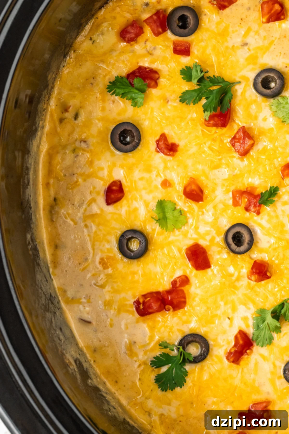 A Crock-Pot filled with a completed taco dip, generously topped with an array of garnishes: melted shredded cheese, vibrant sliced black olives, finely diced tomatoes, and fresh chopped cilantro, making it visually appealing and ready to serve.