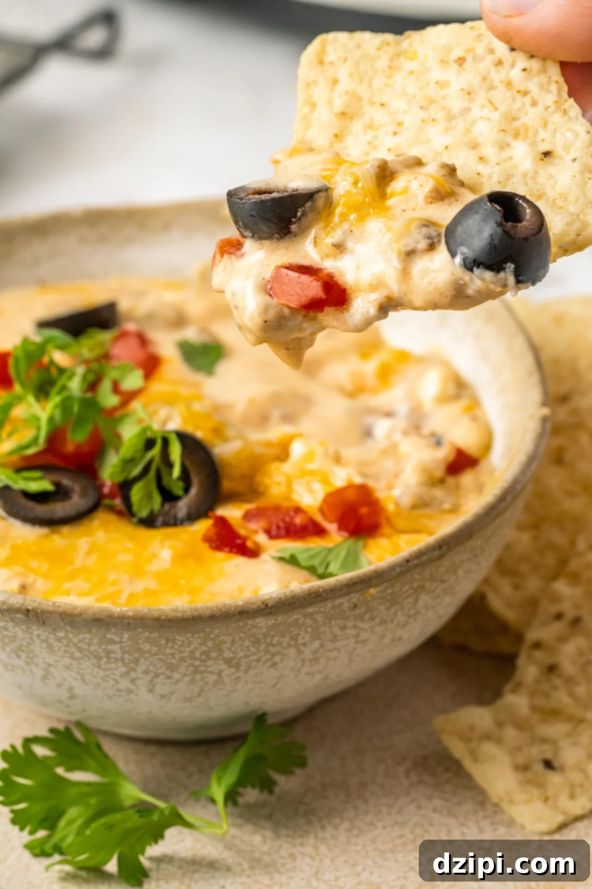 A crisp tortilla chip perfectly dipped into the creamy taco dip, with a small, out-of-focus serving bowl of the dip in the background. It highlights the delicious texture and ideal serving method.