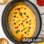 A Crock-Pot filled with a bubbling taco dip, richly topped with melted cheese, vibrant diced tomatoes, thinly sliced black olives, and fresh chopped cilantro. The dip looks hearty and inviting, perfect for scooping.