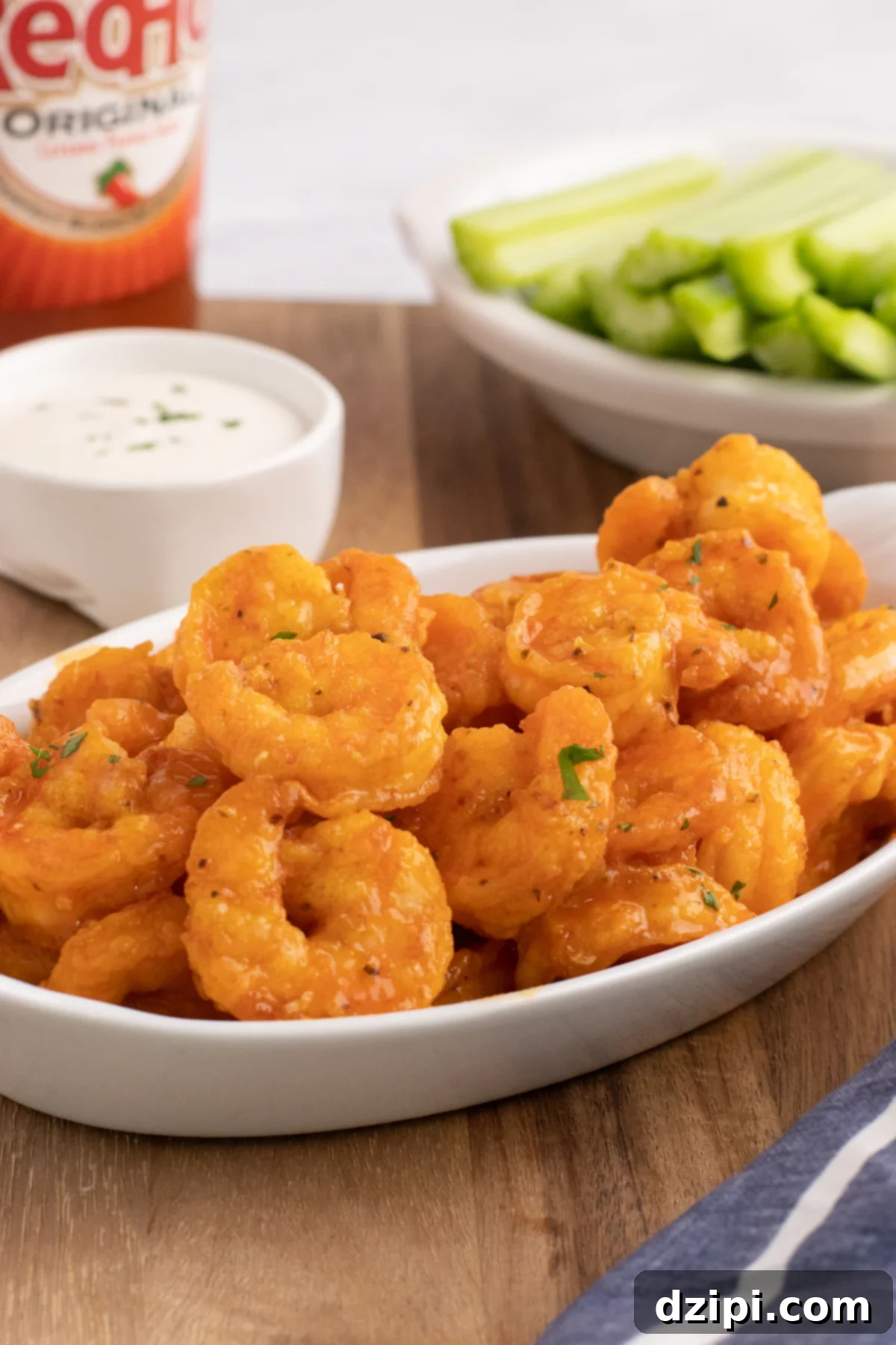A white serving dish filled with buffalo shrimp. Hot sauce, ranch, and celery sticks out of focus in background.