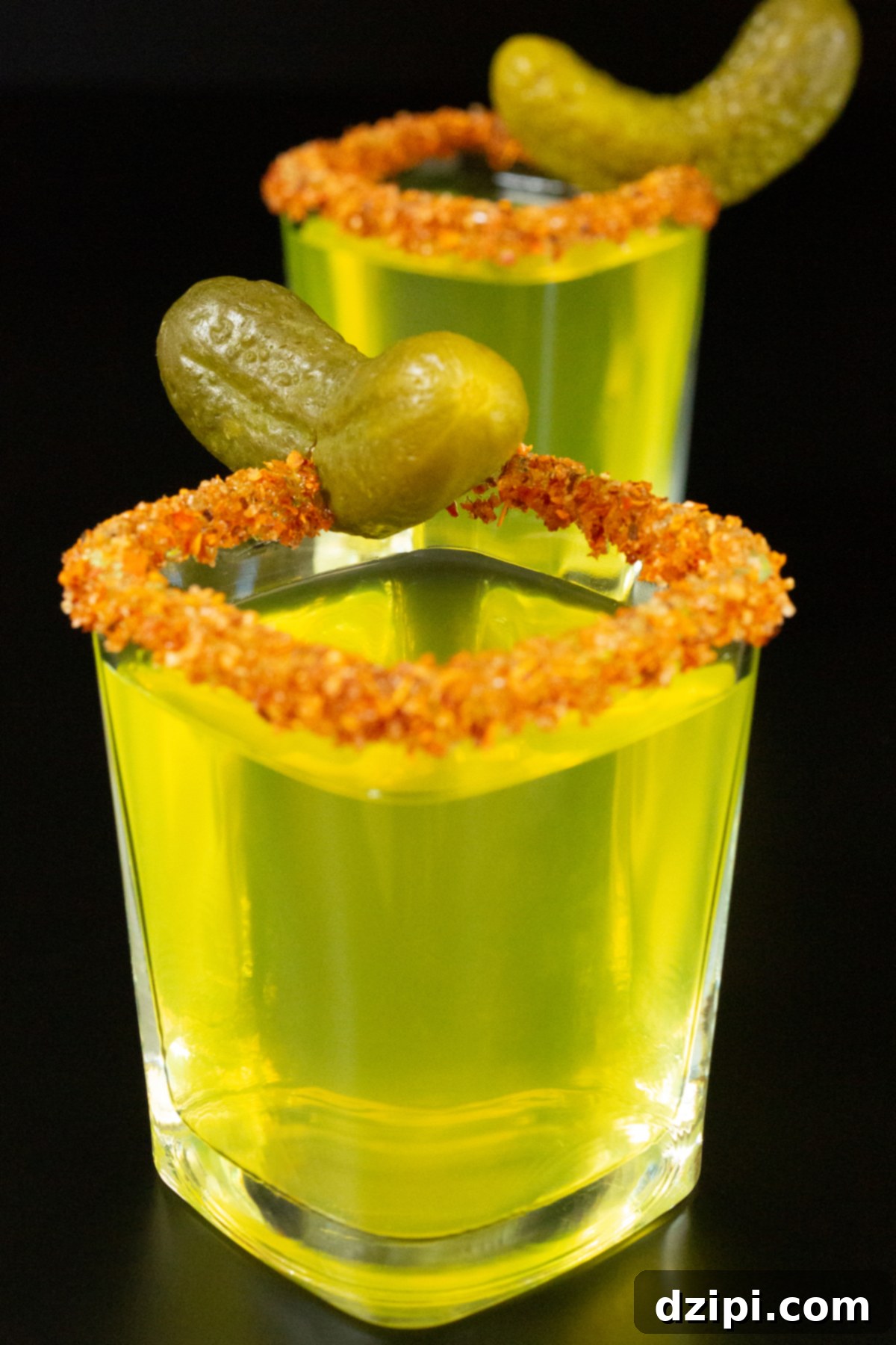 Two square shot glasses filled with neon green pickle shots garnished with a Tajin rim and a baby pickle.