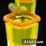 Two square shot glasses filled with neon green pickle shots garnished with a Tajin rim and a baby pickle.